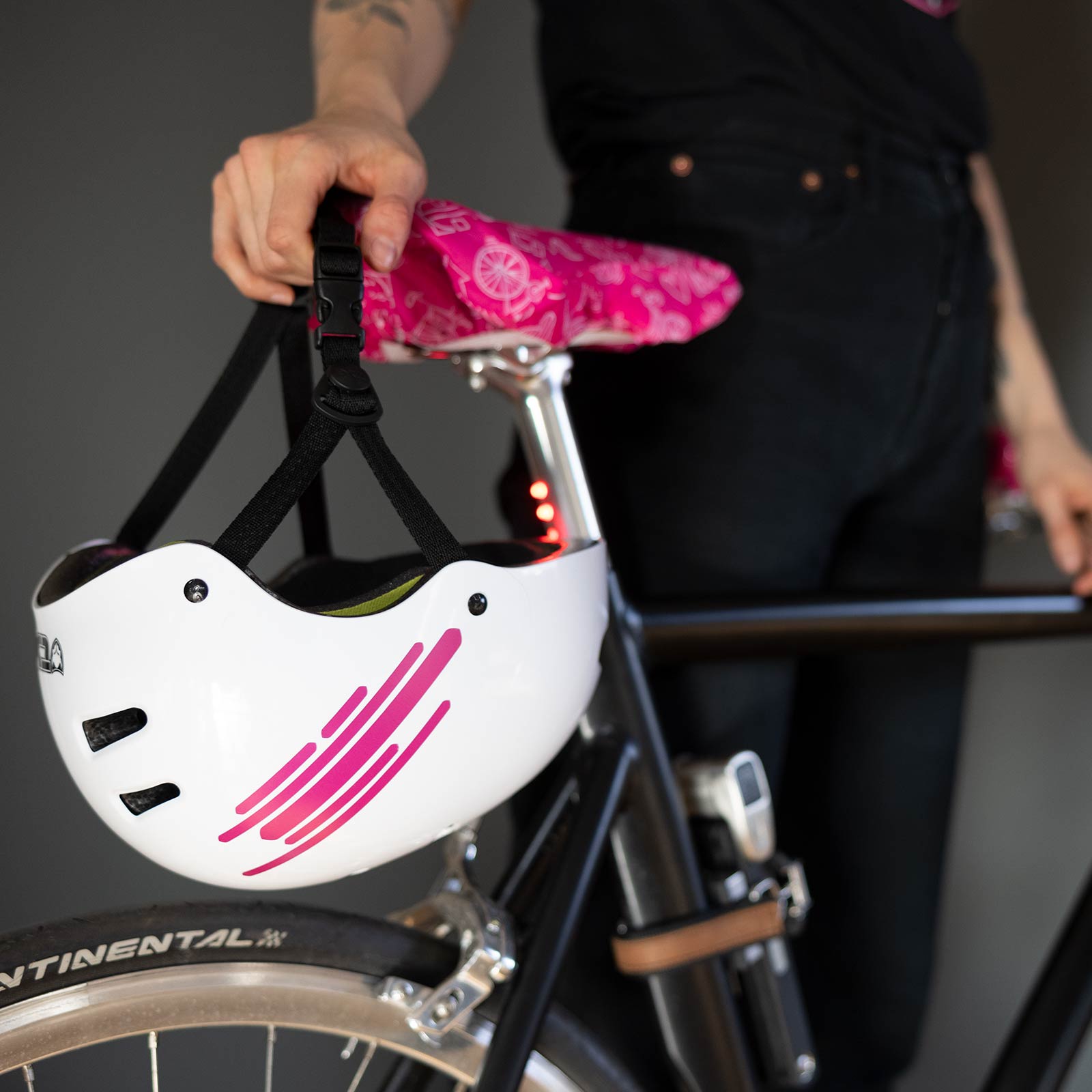Neben einem schwarzen Fahrrad steht eine Person, die einen weißen Helm mit rosa Streifen in der Hand hält. Der Fahrradsattel ist mit einem rosa Bezug mit Mustern bezogen. Der Hintergrund ist unscharf.