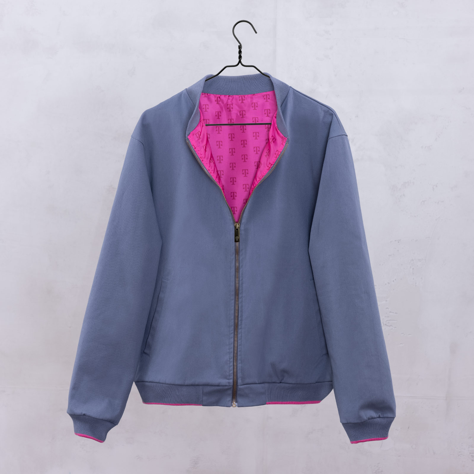 Eine graue Jacke mit Reißverschluss, hellrosa Innenfutter und gemustertem Logo-Aufdruck, die an einem schwarzen Kleiderbügel vor einer hellgrauen Wand hängt.