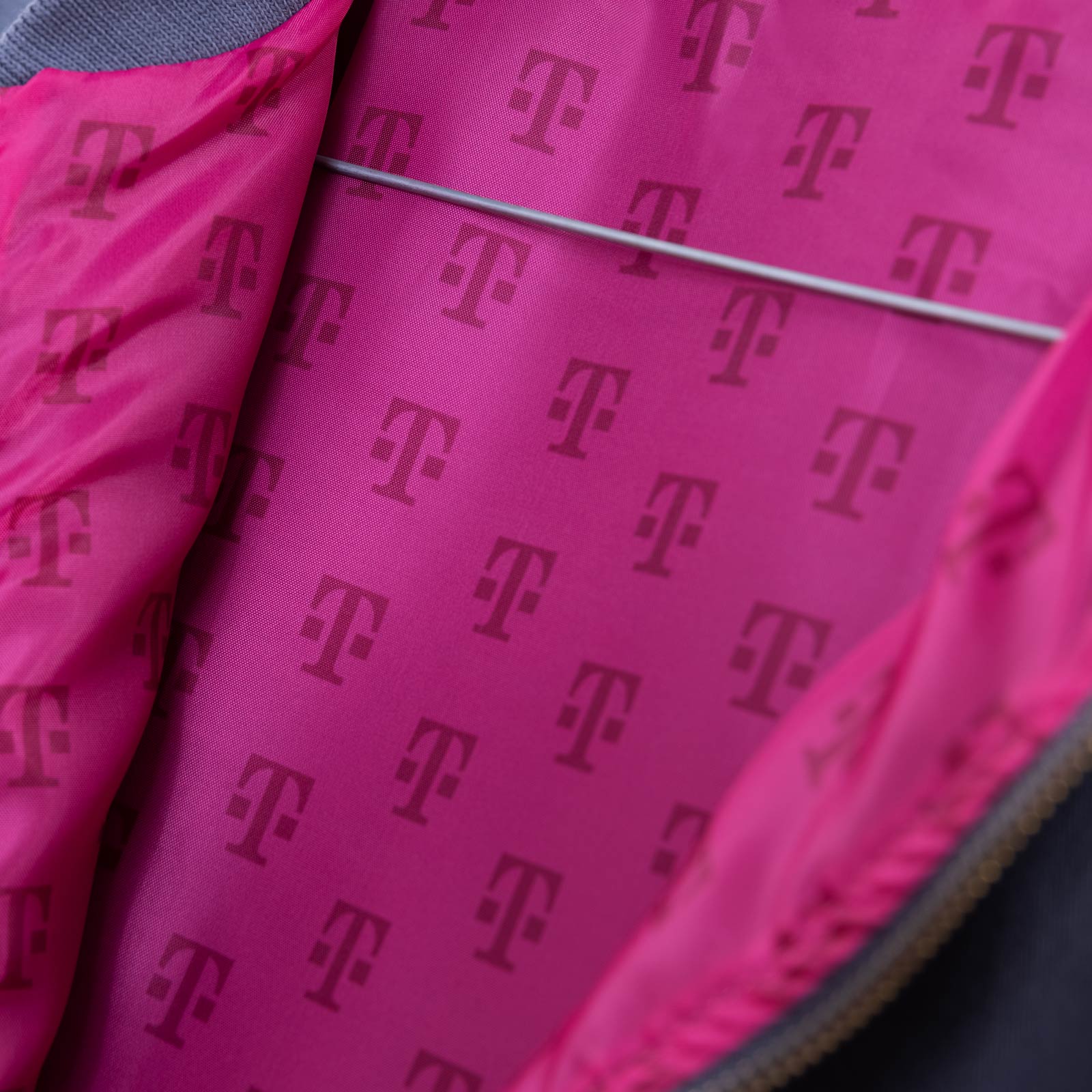 Das Bild zeigt das rosa Innenfutter einer Jacke, das mit sich wiederholenden magentafarbenen T-Logos bedruckt ist. Auch der Rand des Reißverschlusses und ein Metallbügel sind teilweise zu sehen.