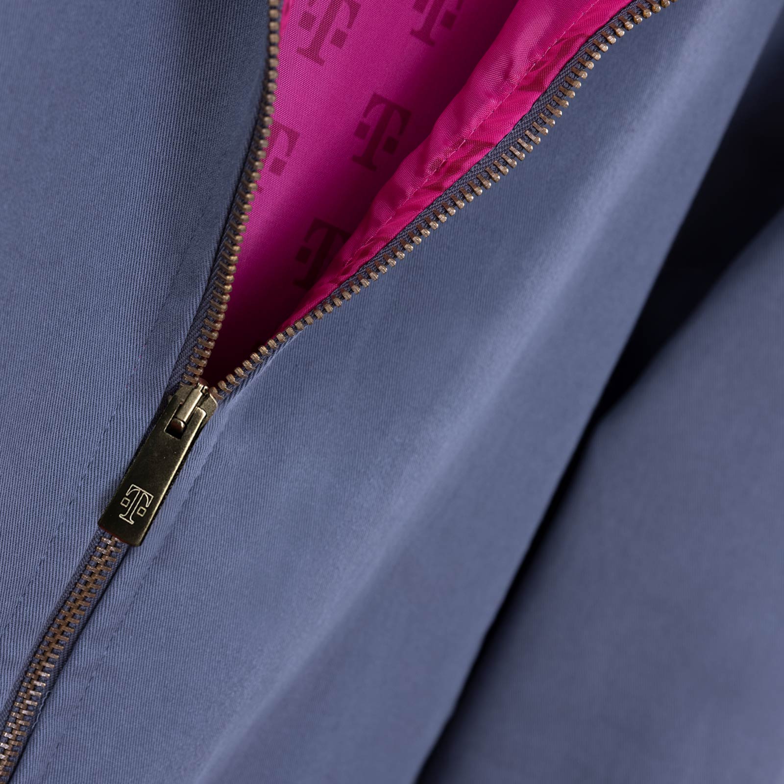 Nahaufnahme einer grauen Jacke mit einem teilweise geöffneten goldenen Reißverschluss, der den Blick auf ein rosa Innenfutter freigibt, das mit sich wiederholenden T-Logos bedruckt ist.