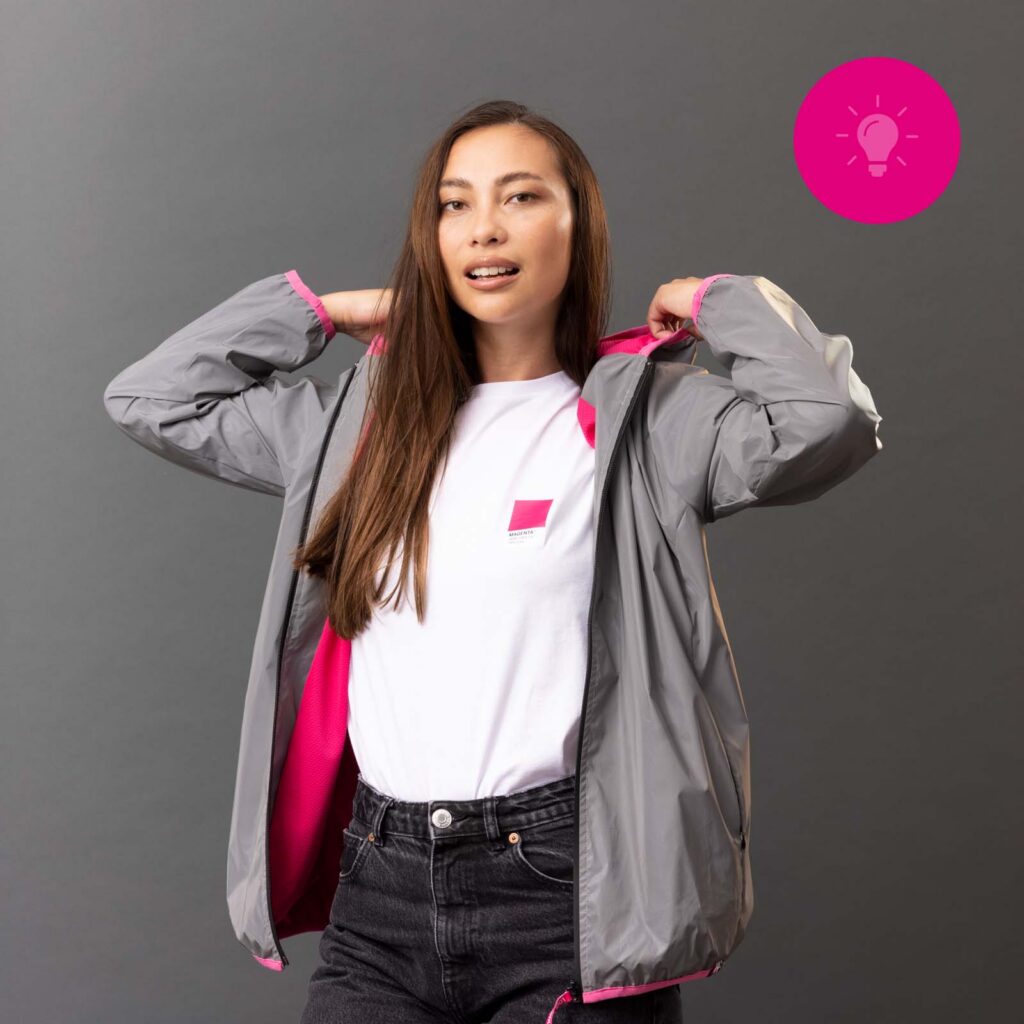 Eine Frau in einer grauen Jacke mit rosa Futter steht vor einem grauen Hintergrund und hält die Jacke offen. Sie trägt ein weißes Hemd und schwarze Jeans. In der oberen rechten Ecke ist das Symbol einer rosa Glühbirne zu sehen.