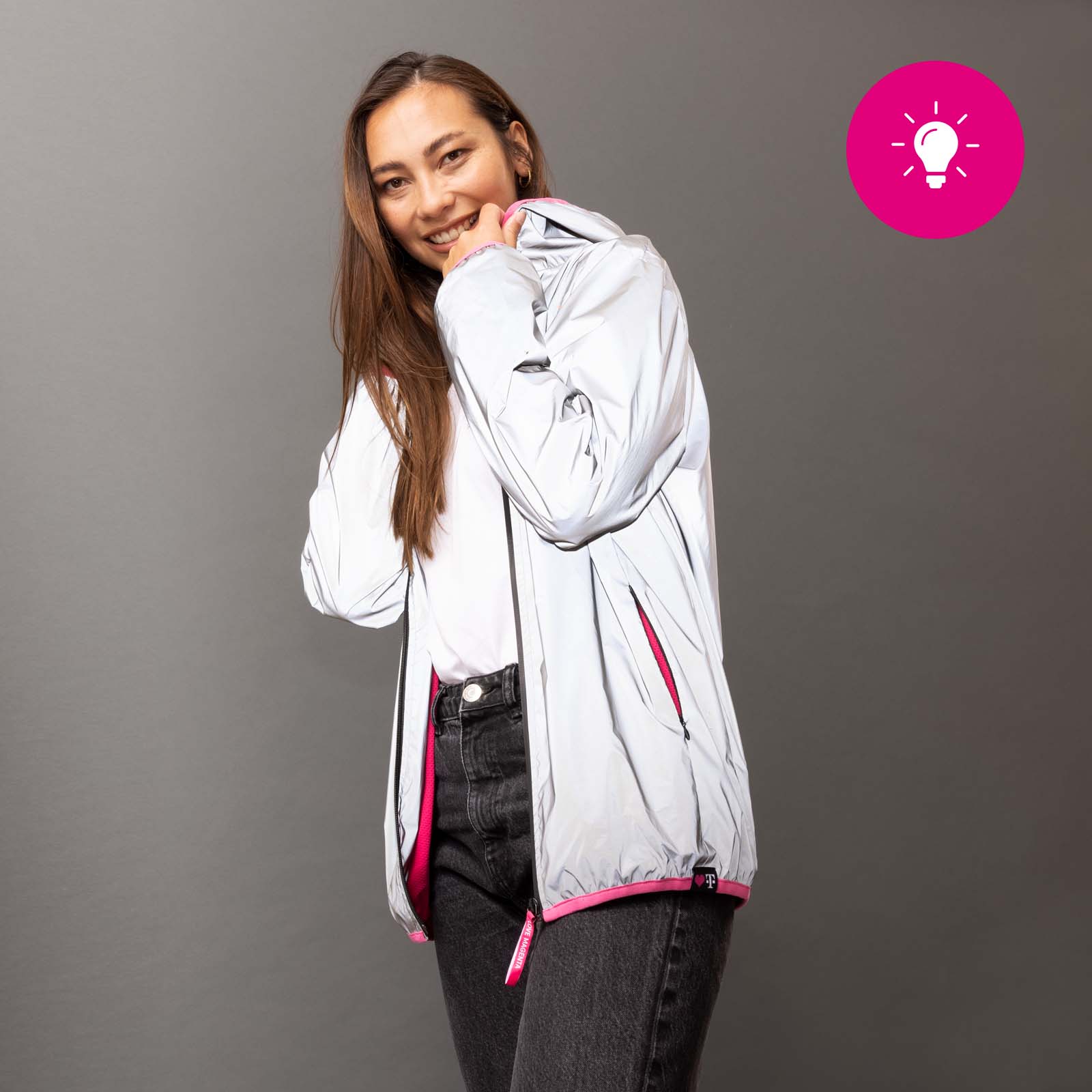 Eine lächelnde Frau, die eine hellgraue Jacke mit rosa Akzenten über einem weißen Hemd und einer dunklen Jeans trägt, steht vor einem grauen Hintergrund. In der oberen rechten Ecke befindet sich ein rosa Kreis mit einem Glühbirnen-Symbol.