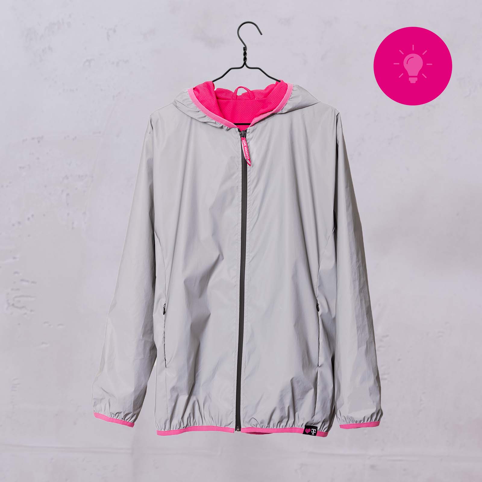 Eine graue Jacke mit rosa Innenfutter und Akzenten hängt auf einem schwarzen Kleiderbügel vor einem hellen Hintergrund. In der rechten oberen Ecke ist ein rosa Kreis mit einem Glühbirnen-Symbol zu sehen.
