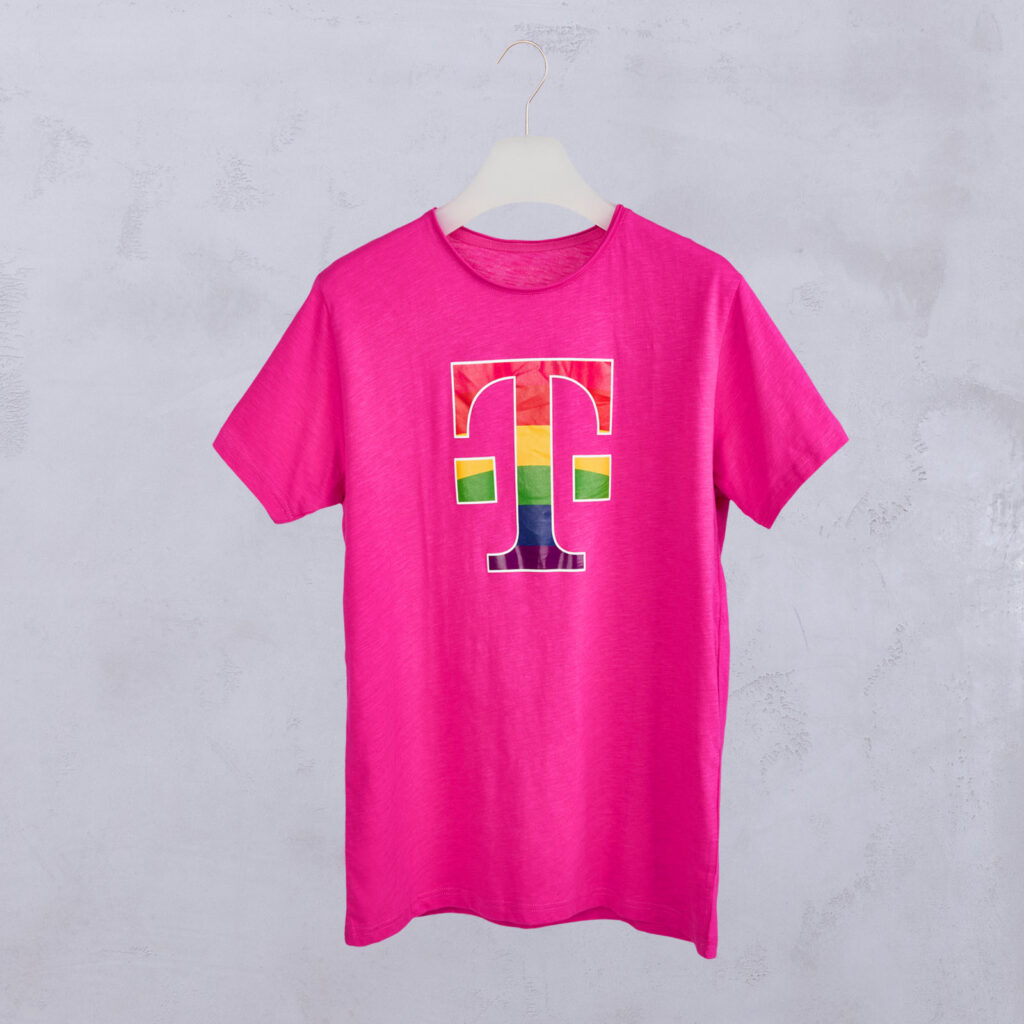 Ein leuchtend pinkes T-Shirt mit einem großen Buchstaben „T“ auf der Vorderseite, der mit einem Regenbogen-Farbverlauf ausgefüllt ist. Das Shirt hängt auf einem weißen Kleiderbügel vor einer hellgrauen, strukturierten Wand.