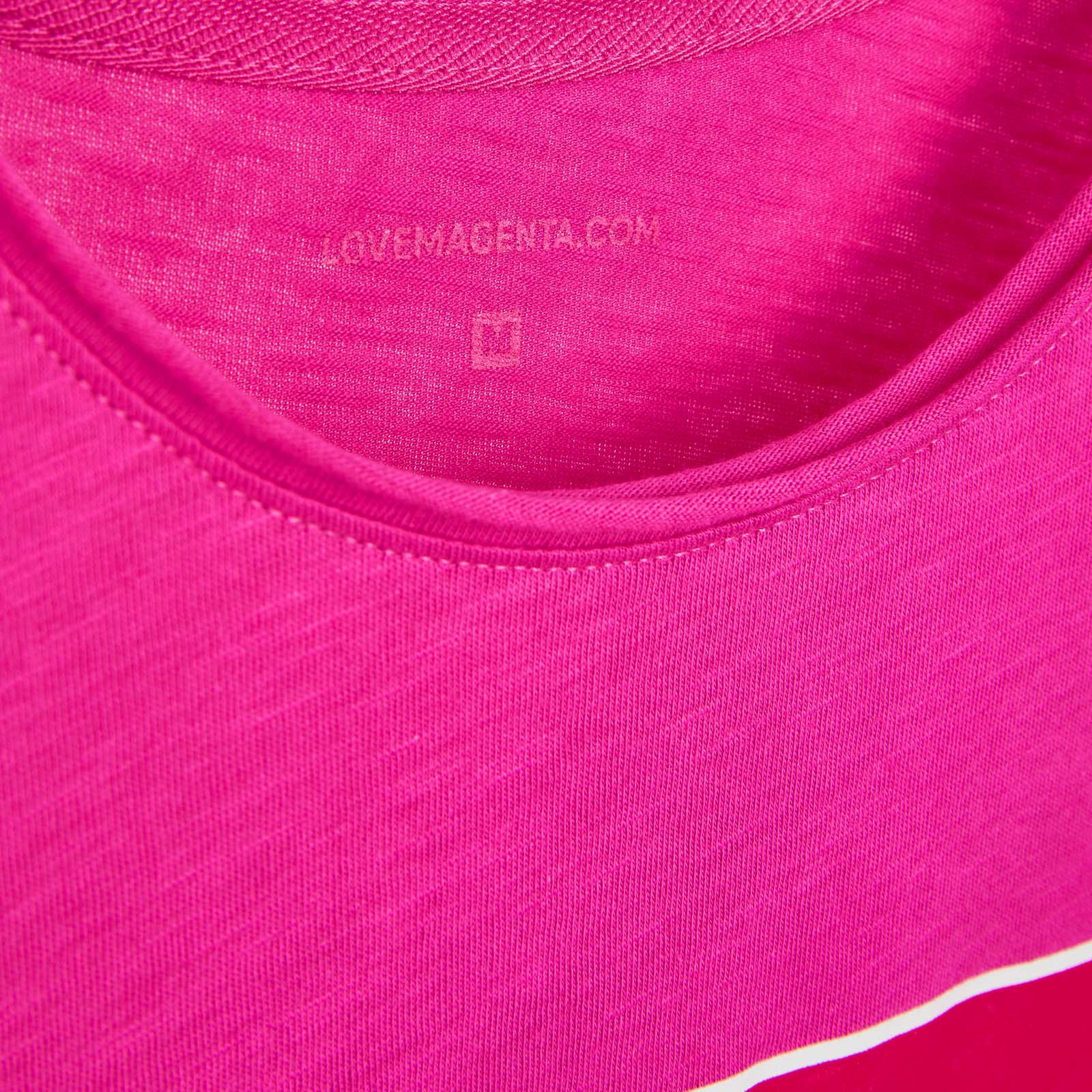 Nahaufnahme des Ausschnitts eines magentafarbenen Shirts, die die Stoffstruktur und ein auf der Innenseite aufgedrucktes Etikett mit der Aufschrift „LOVEMAGENTA.COM“ und einem kleinen quadratischen Symbol darunter zeigt.