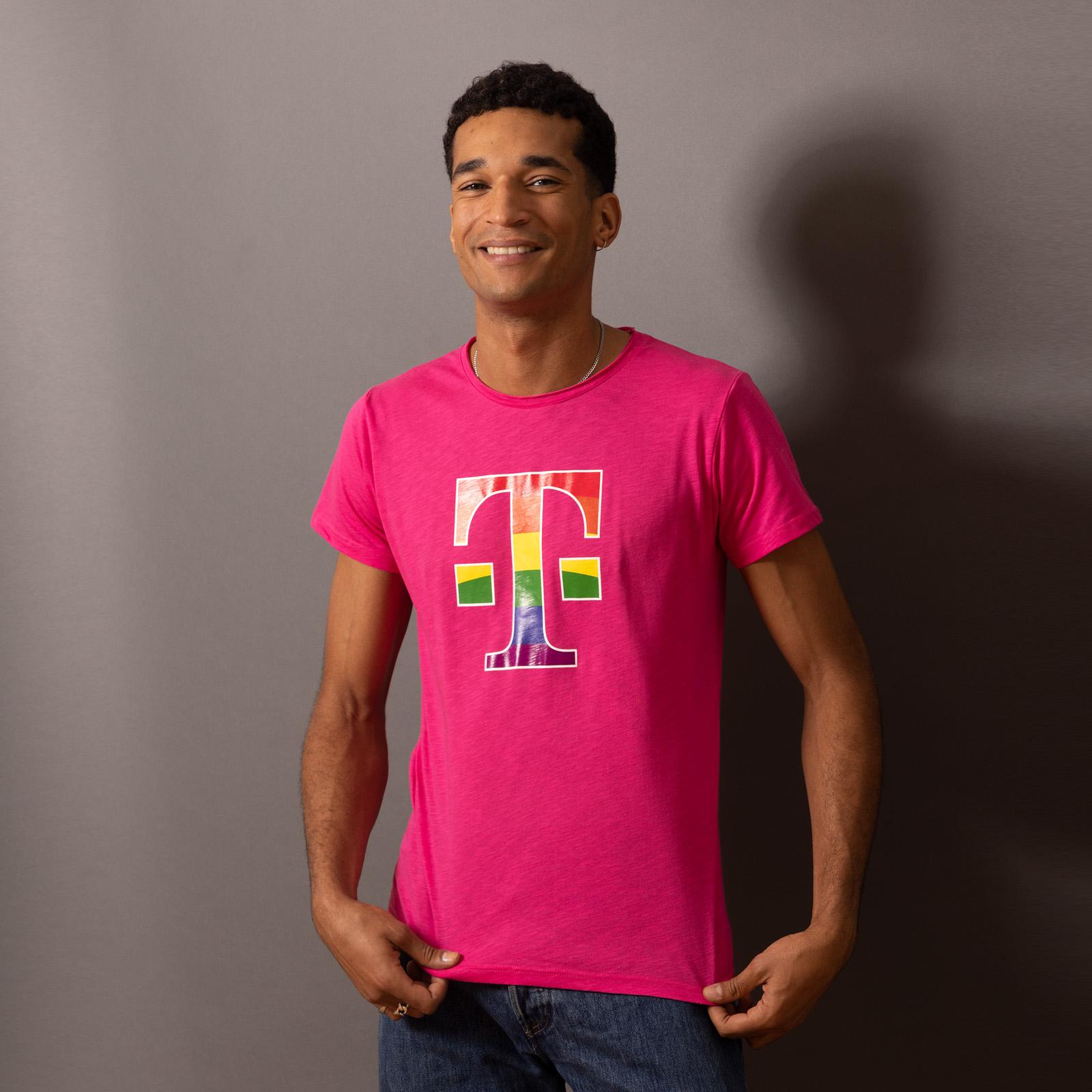 Eine Person steht vor einer grauen Wand, lächelt und trägt ein leuchtend pinkes T-Shirt, auf dessen Vorderseite ein großes, stilisiertes „T“ und bunte Formen aufgedruckt sind.