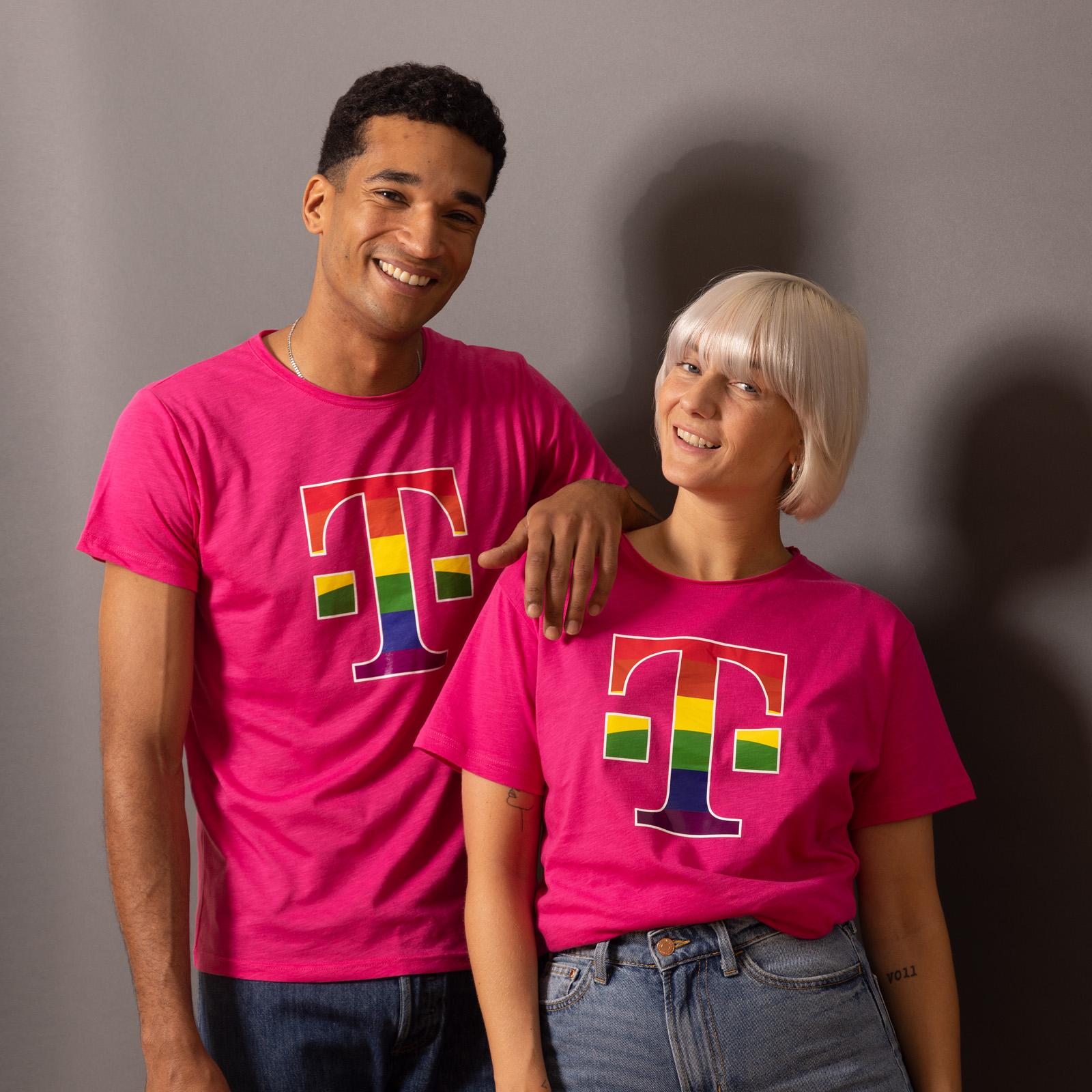 Zwei Personen lächeln in die Kamera; sie tragen passende rosa T-Shirts mit einem regenbogenfarbenen T-Logo und stehen vor einem grauen Hintergrund.