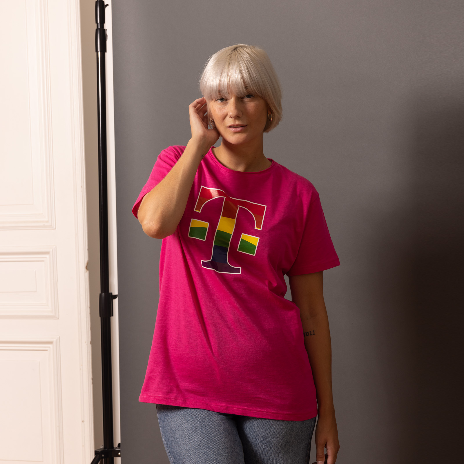 Eine Person mit kurzen blonden Haaren trägt ein leuchtend pinkes T-Shirt mit einem großen, bunten T-Logo und eine blaue Jeans und steht vor einem grauen Hintergrund und einer weißen Tür.