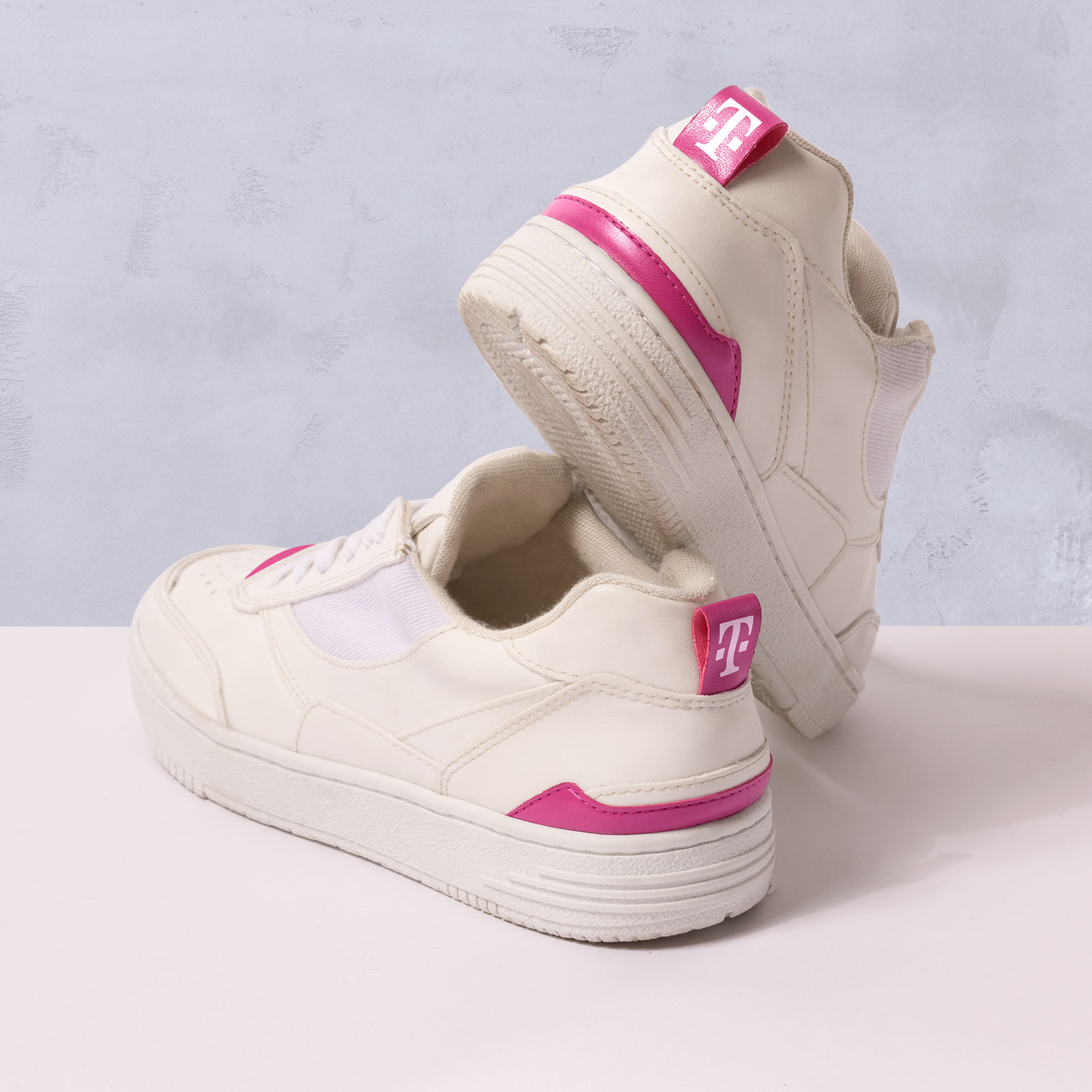 Zu sehen sind zwei weiße Sneaker mit rosa Akzenten und einem T-Logo an der Ferse: Der eine steht aufrecht, der andere lehnt daran, vor einem hellen Hintergrund.