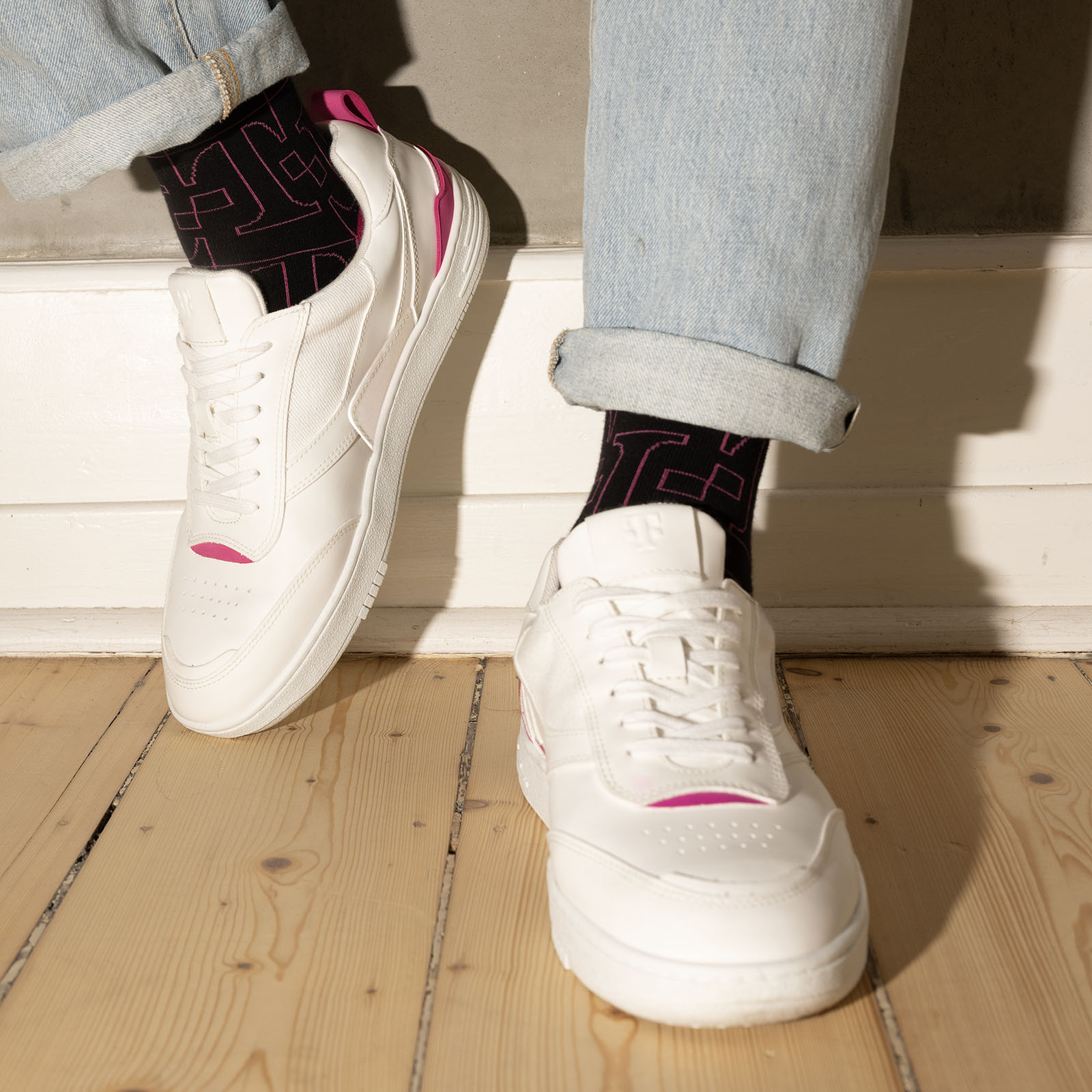 Eine Person, die hellblaue Jeans, weiße Sneaker mit rosa Akzenten und schwarze Socken mit geometrischem Muster trägt, steht auf einem hellen Holzboden in der Nähe einer weißen Wand.