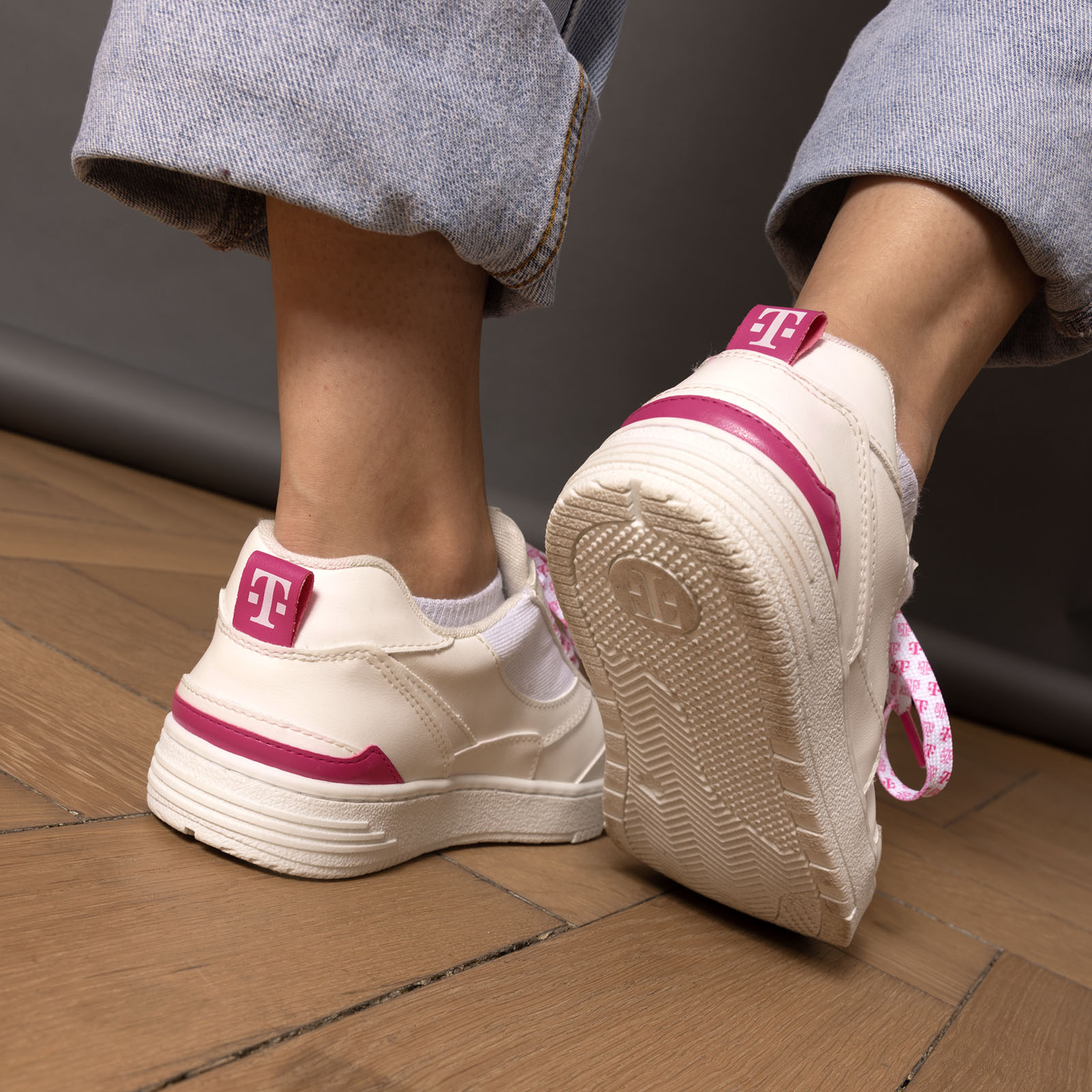 Eine Person, die helle Sneaker mit rosa Akzenten und eine blaue Jeans mit Umschlag trägt und auf einem Holzboden steht. Der Fokus liegt auf den Schuhen, wobei die Sohle und ein rosa Logo an der Ferse zu sehen sind.