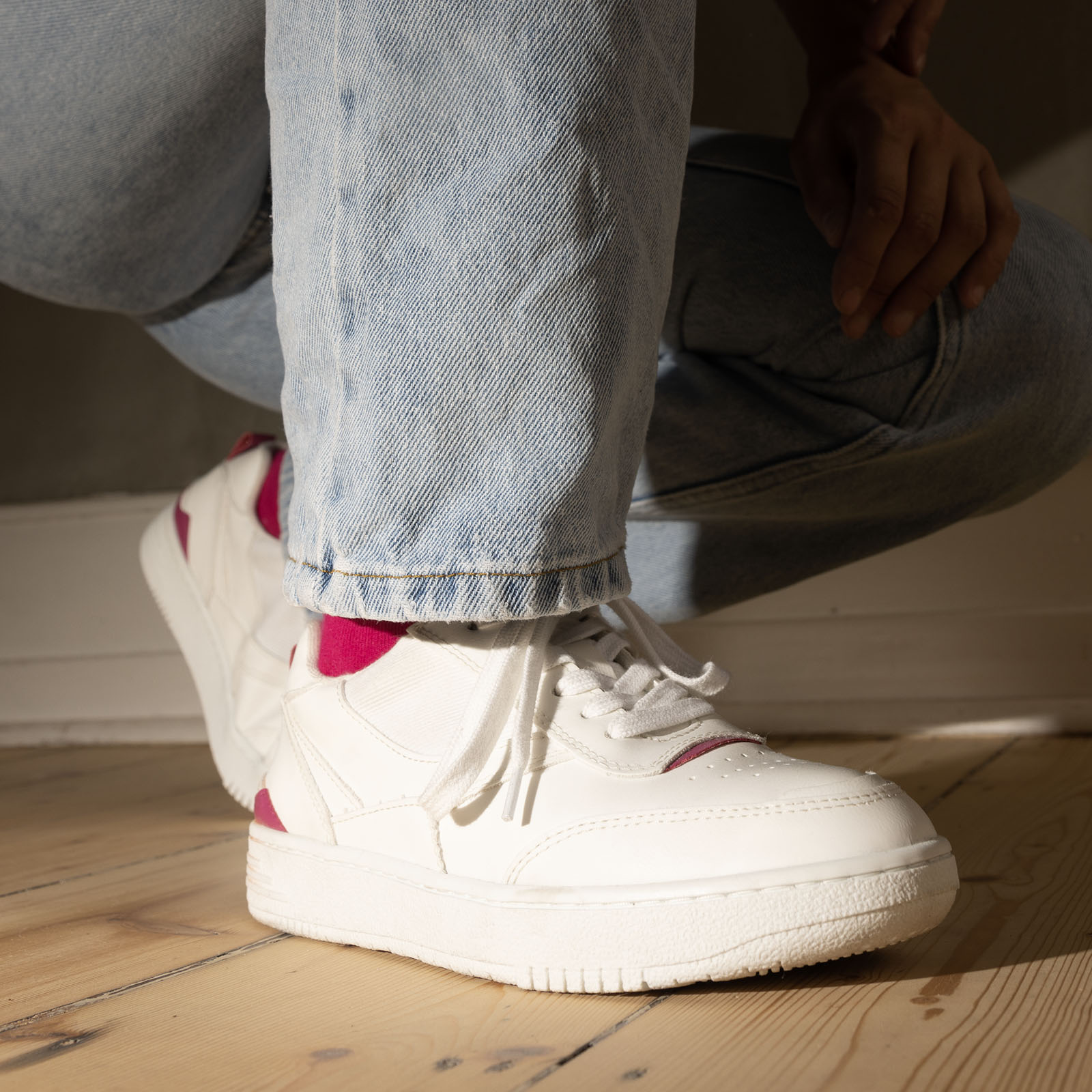 Eine Person, die hellblaue Jeans und weiße Turnschuhe mit rosa Akzenten trägt, hockt auf einem hellen Holzboden, während das Sonnenlicht Schatten auf den Boden wirft.
