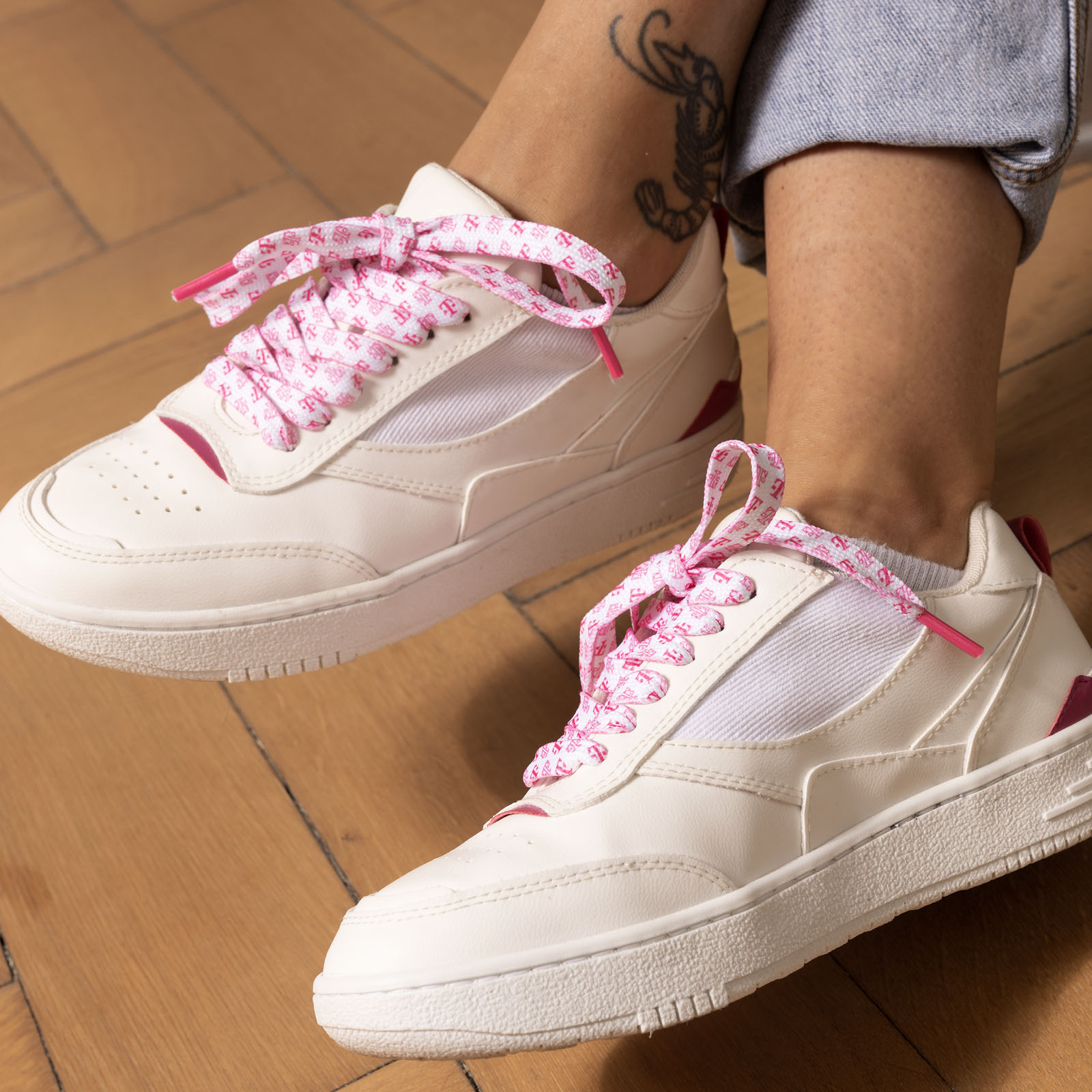 Weiße Sneaker mit rosa Details und gemusterten rosa Schnürsenkeln, getragen von einer Person mit einem Tattoo am Knöchel. Im Hintergrund ist ein Holzboden zu sehen.