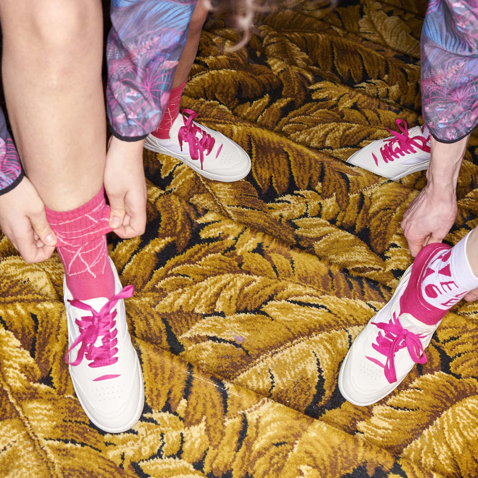 Zwei Personen binden die Schnürsenkel ihrer weißen Turnschuhe mit rosa Schnürsenkeln zusammen und tragen rosa Socken. Sie stehen auf einem Teppich mit einem auffälligen gelben Blattmuster. Nur ihre Beine und Hände sind zu sehen.