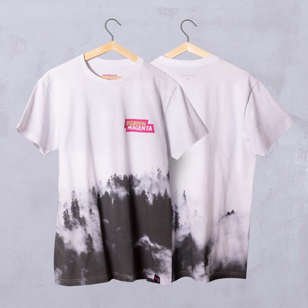 Auf zwei weißen T-Shirts, die an Kleiderbügeln hängen, ist am unteren Rand ein schwarz-graues Motiv mit Wald und Nebel zu sehen. Auf der Vorderseite befindet sich auf Brusthöhe ein rosa #GREEN MAGENTA-Etikett. Beide Shirts hängen vor einer hellgrauen Wand.