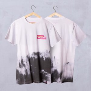 Auf zwei weißen T-Shirts, die an Kleiderbügeln hängen, ist am unteren Rand ein schwarz-graues Motiv mit Wald und Nebel zu sehen. Auf der Vorderseite befindet sich auf Brusthöhe ein rosa #GREEN MAGENTA-Etikett. Beide Shirts hängen vor einer hellgrauen Wand.