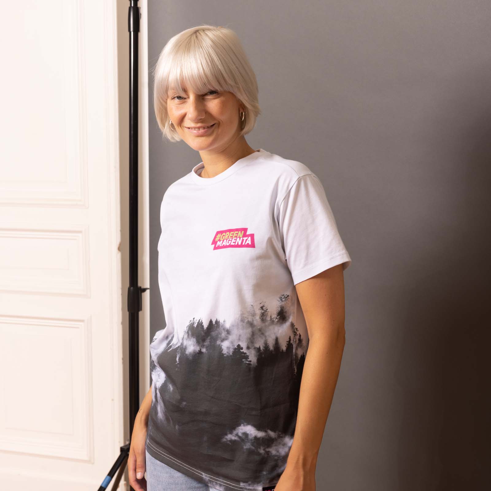 Eine Frau mit kurzen blonden Haaren lächelt; sie trägt ein weißes T-Shirt mit einem Waldmotiv und einem rosa MODERN MAGENTA-Logo und steht in einem Innenraum vor einem grau-weißen Hintergrund.
