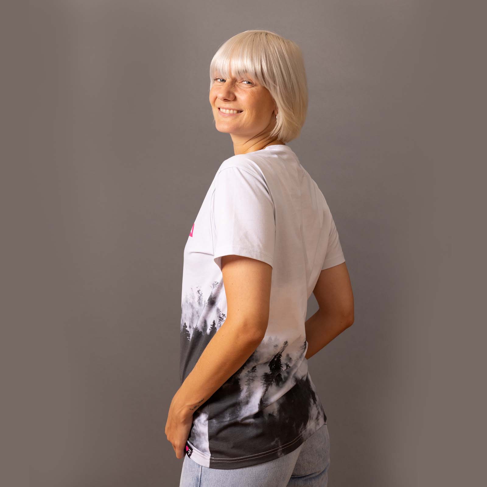 Eine Person mit kurzen blonden Haaren steht seitlich zur Kamera, lächelt in die Kamera und trägt ein schwarz-weißes Tie-Dye-T-Shirt sowie hellblaue Jeans vor einem schlichten grauen Hintergrund.