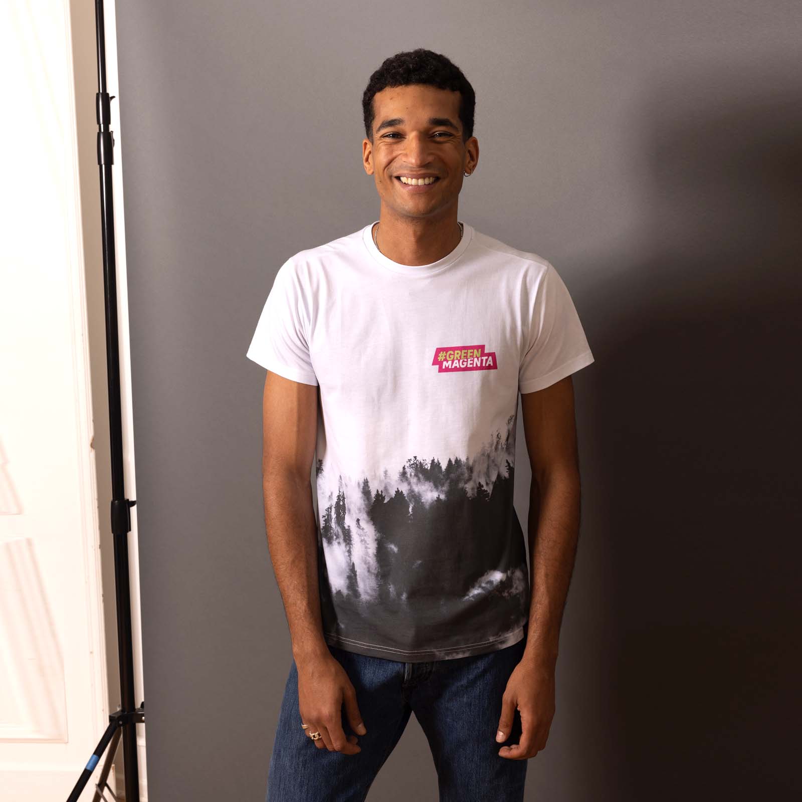Eine Person, die vor einer grauen Wand steht und lächelt; sie trägt ein schwarz-weißes T-Shirt mit einem rot-weißen Logo und eine blaue Jeans.