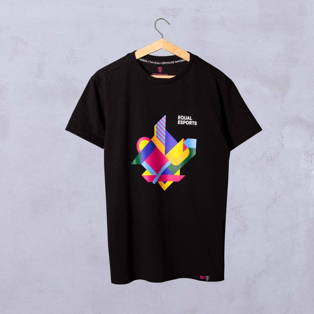 Auf einem Kleiderbügel hängt ein schwarzes T-Shirt mit einem bunten geometrischen Muster und der Aufschrift „EQUAL ESPORTS“ auf der Brust. Im Hintergrund ist eine hellgraue Wand zu sehen.