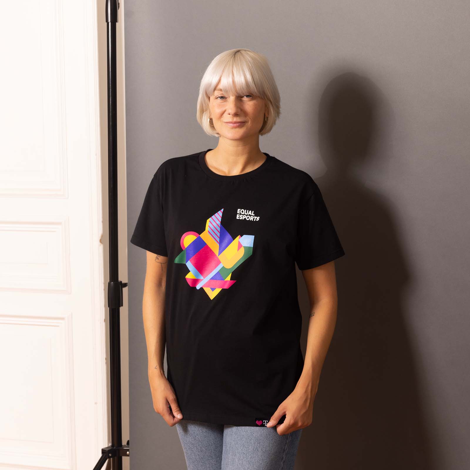 Eine Person mit kurzen weißen Haaren steht vor einer grauen Wand und trägt ein schwarzes T-Shirt mit einem bunten Herzmotiv und der Aufschrift „Equal Esports“.