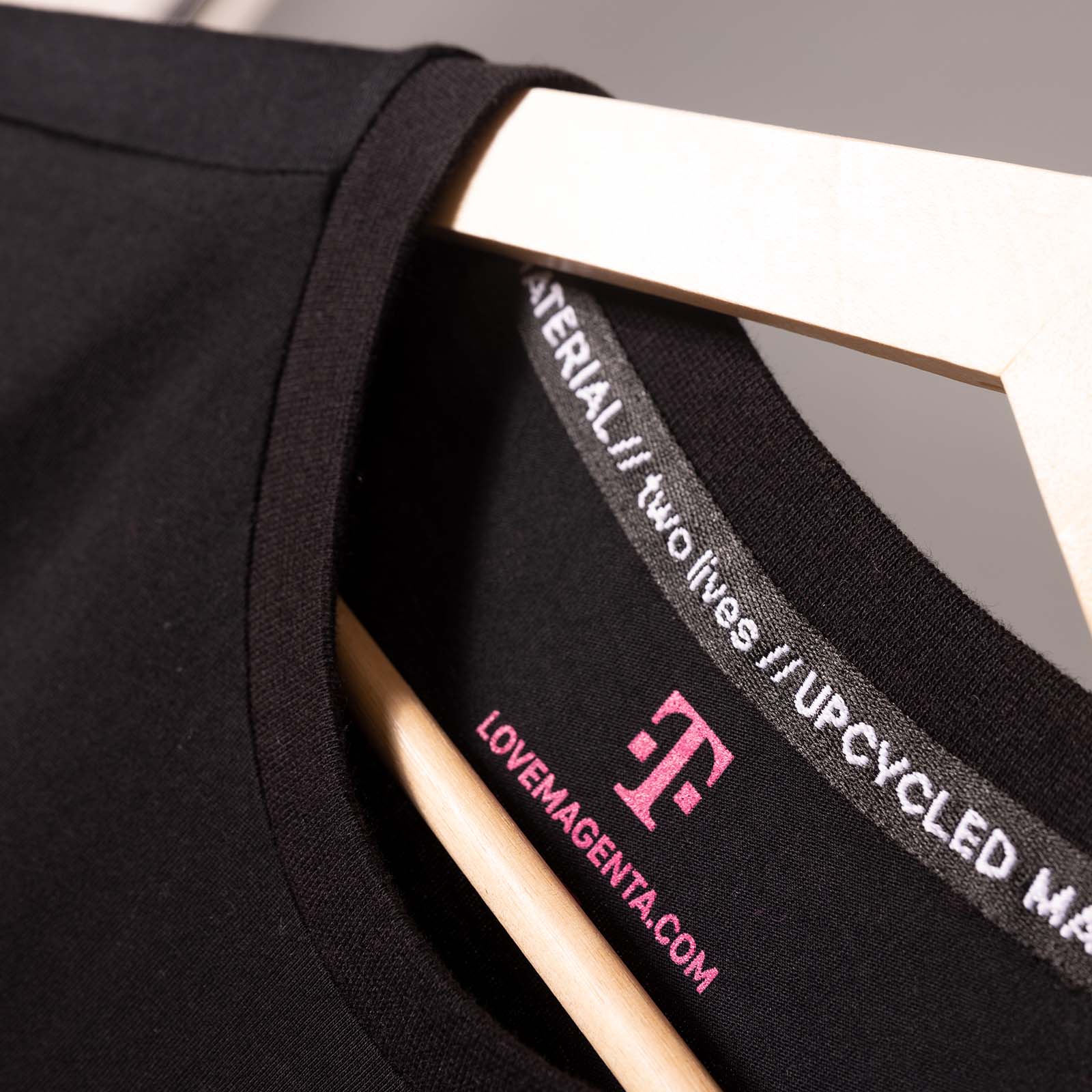 Nahaufnahme eines schwarzen T-Shirts auf einem Kleiderbügel, auf der der Kragen und das Innenetikett zu sehen sind. Auf dem Etikett steht „LOVEMAGENTA.COM“ mit einem rosa Logo; außerdem sind die Angaben „UPCYCLED MATERIAL“ und „two lives“ am inneren Halsausschnitt zu lesen.