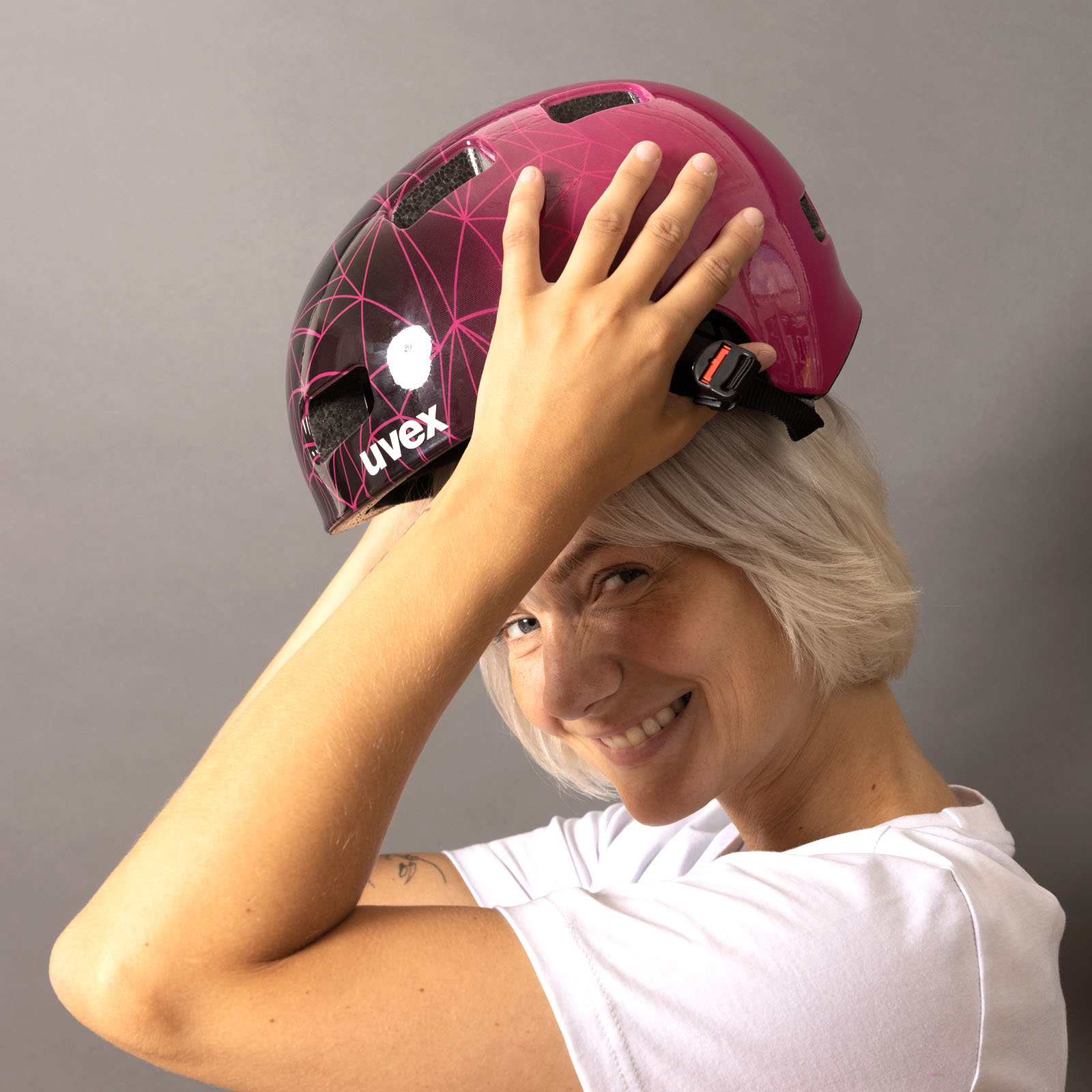 Eine lächelnde Person mit kurzen, hellen Haaren rückt sich einen rosa-schwarzen Fahrradhelm auf dem Kopf zurecht; sie trägt ein weißes Hemd und steht vor einem schlichten grauen Hintergrund.