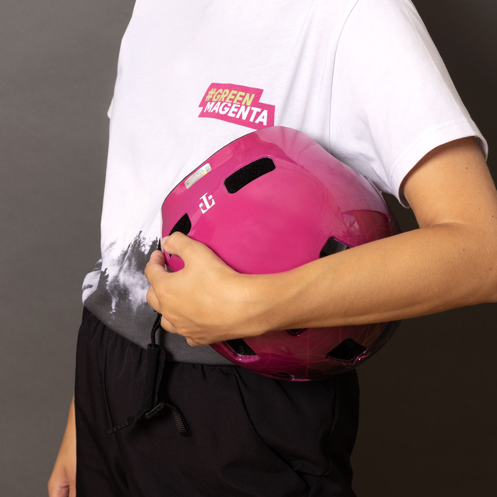 Vor einem schlichten Hintergrund hält eine Person in einem weißen #GREENMAGENTA-Shirt einen leuchtend pinkfarbenen Helm unter dem Arm.
