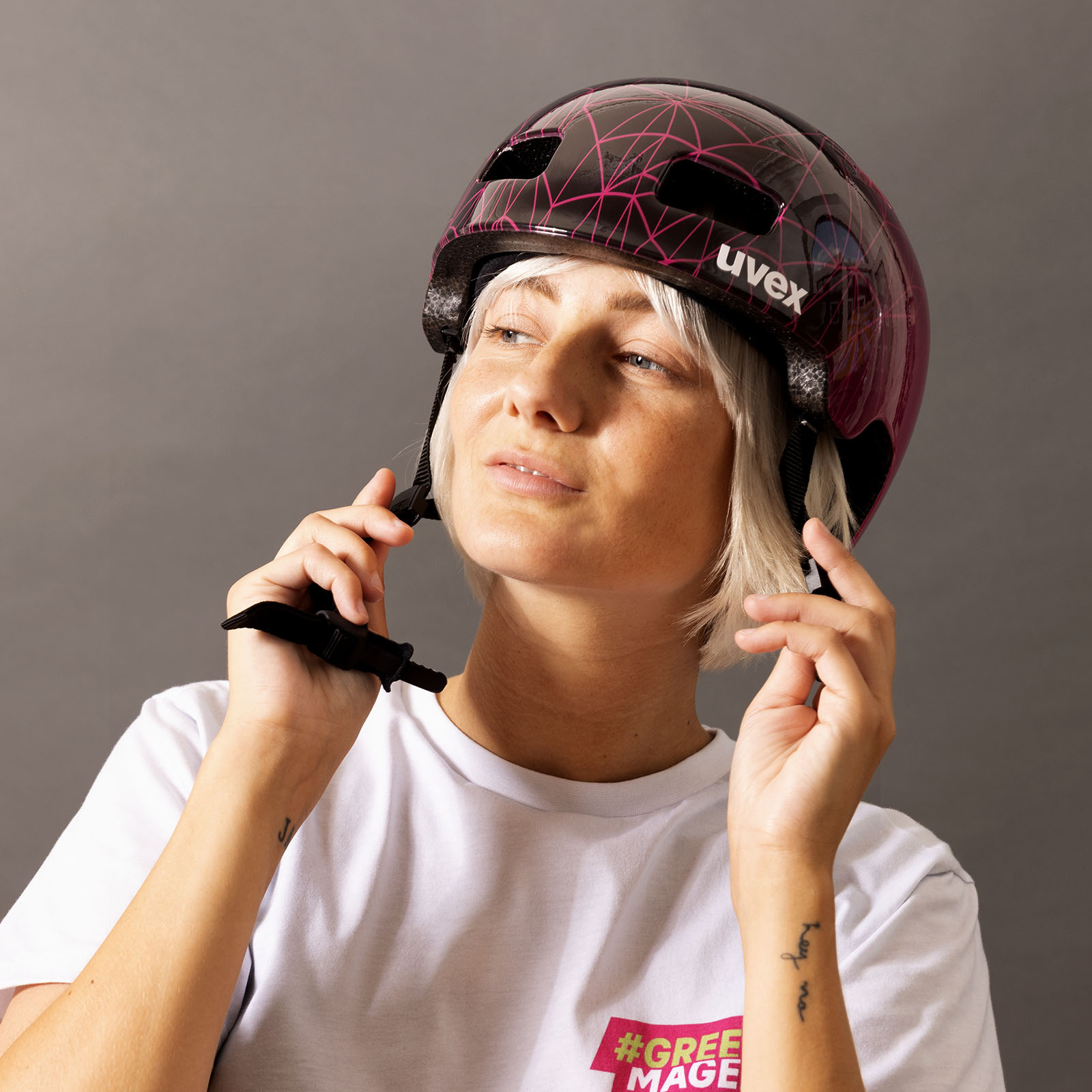 Eine Person mit kurzen blonden Haaren rückt einen lila-schwarzen UVEX-Helm zurecht. Sie trägt ein weißes T-Shirt mit einem rosa-gelben Aufdruck und steht vor einem einfarbigen grauen Hintergrund.