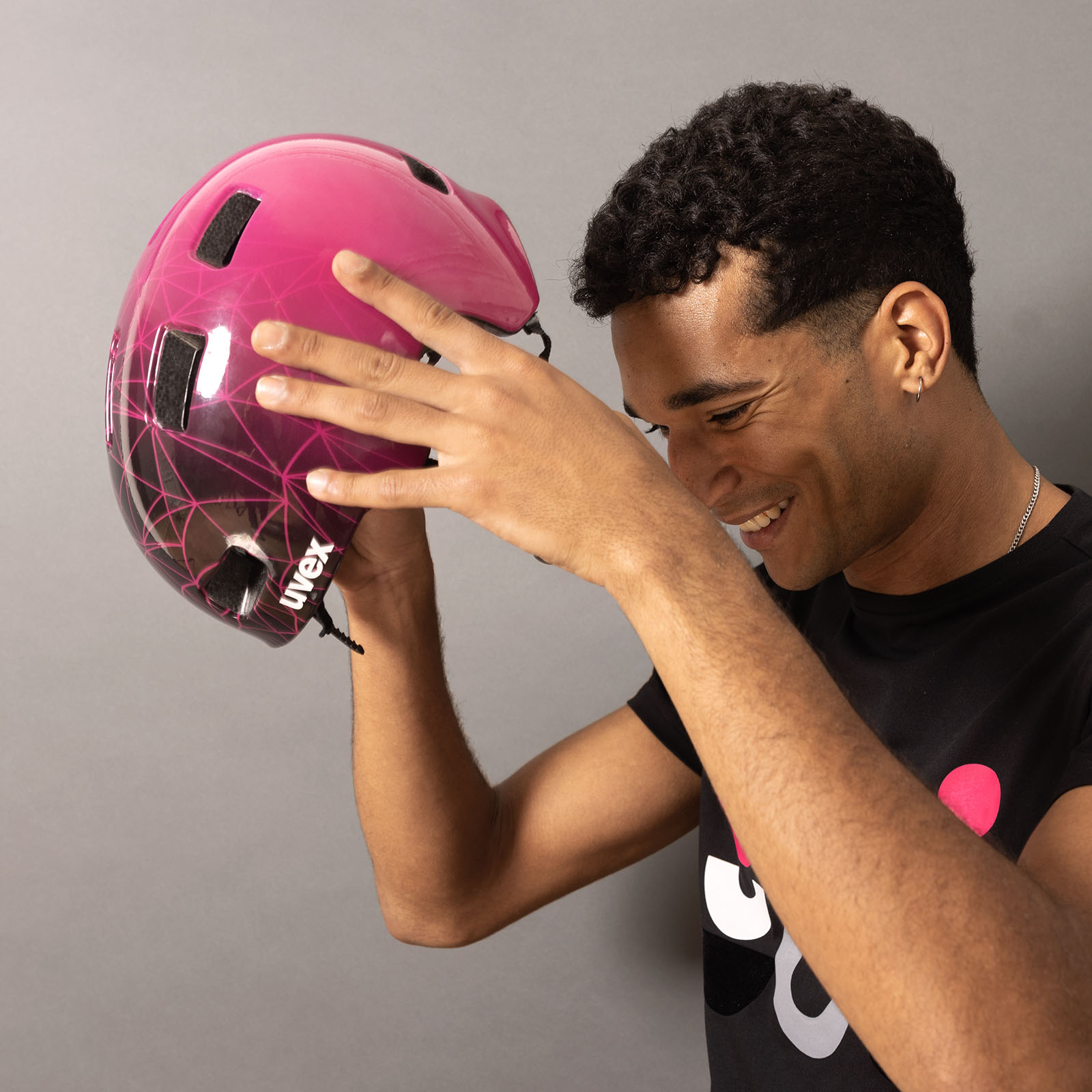 Eine lächelnde Person mit kurzen Locken hält einen rosa Helm über den Kopf und macht sich bereit, ihn aufzusetzen. Sie trägt ein schwarzes T-Shirt mit einem bunten, abstrakten Muster und steht vor einem schlichten grauen Hintergrund.