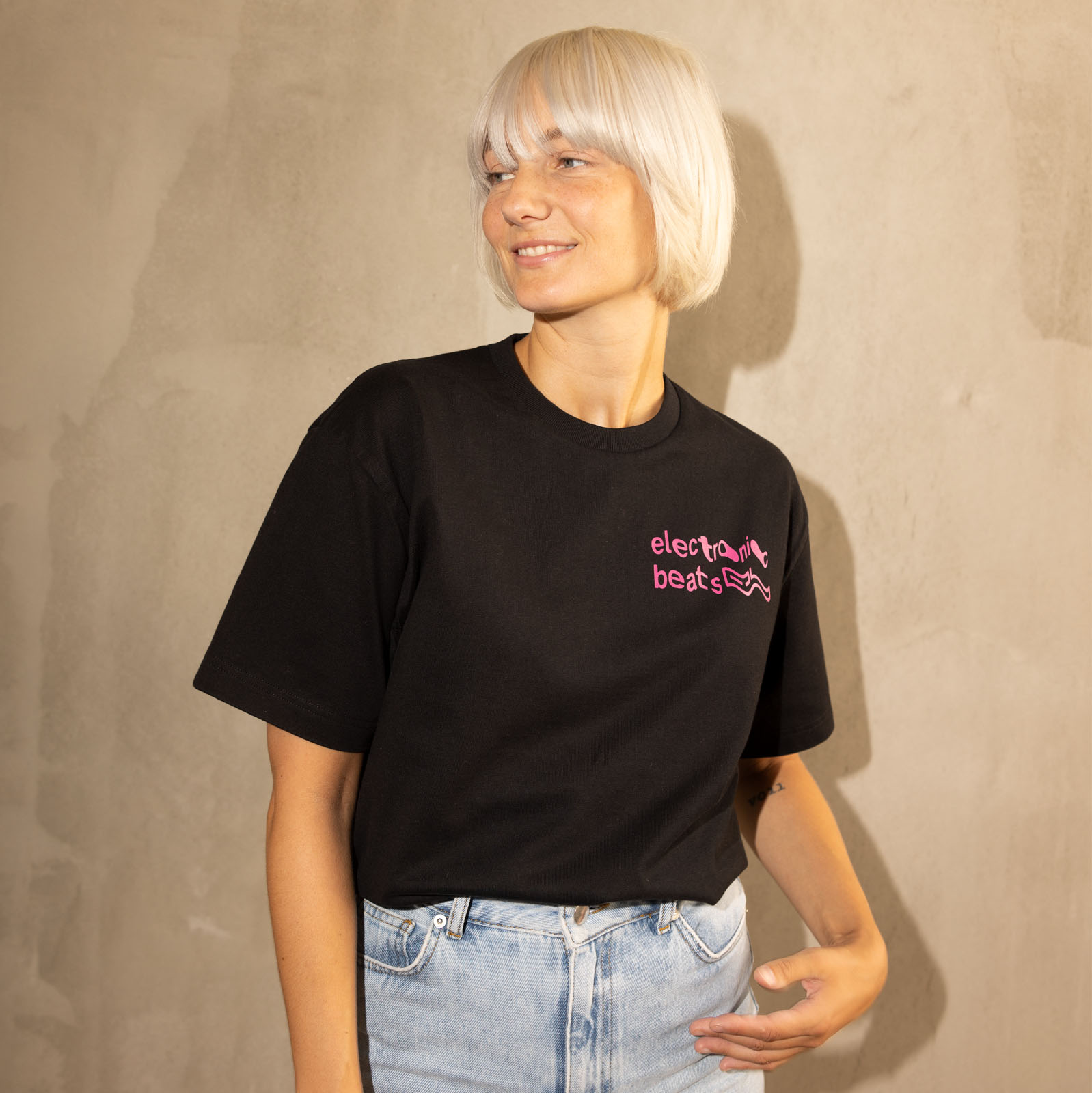 Eine Frau mit kurzen, platinblonden Haaren, die ein schwarzes „Electronic Beats“-T-Shirt und hellblaue Jeans trägt, steht vor einer beigen Wand und blickt mit einem leichten Lächeln zur Seite.