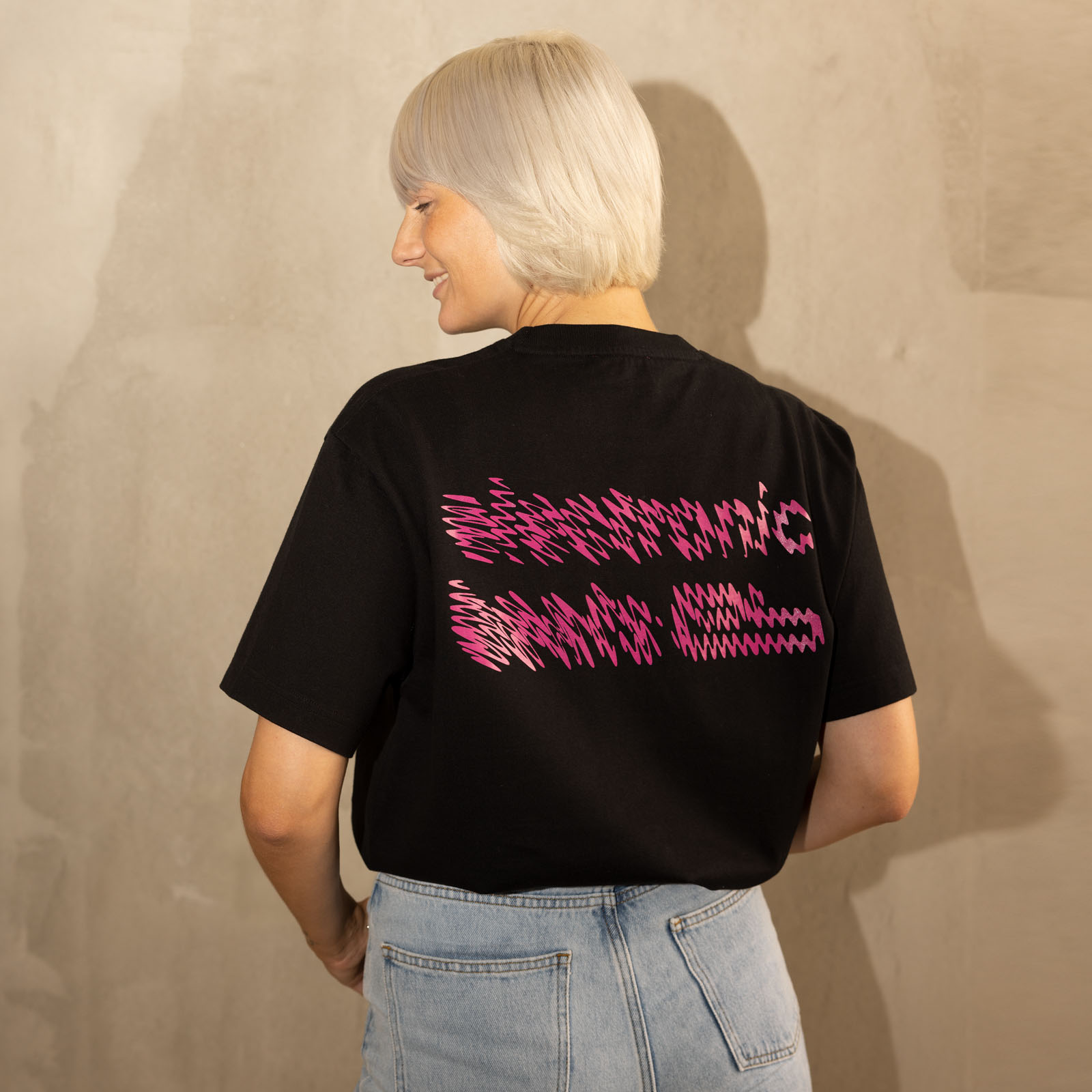 Eine Person mit kurzem, platinblondem Haar steht mit dem Rücken zur Kamera, trägt ein schwarzes T-Shirt mit rosa, wellenförmigem Schriftzug und hellblaue Jeans und steht vor einer beigen, strukturierten Wand.