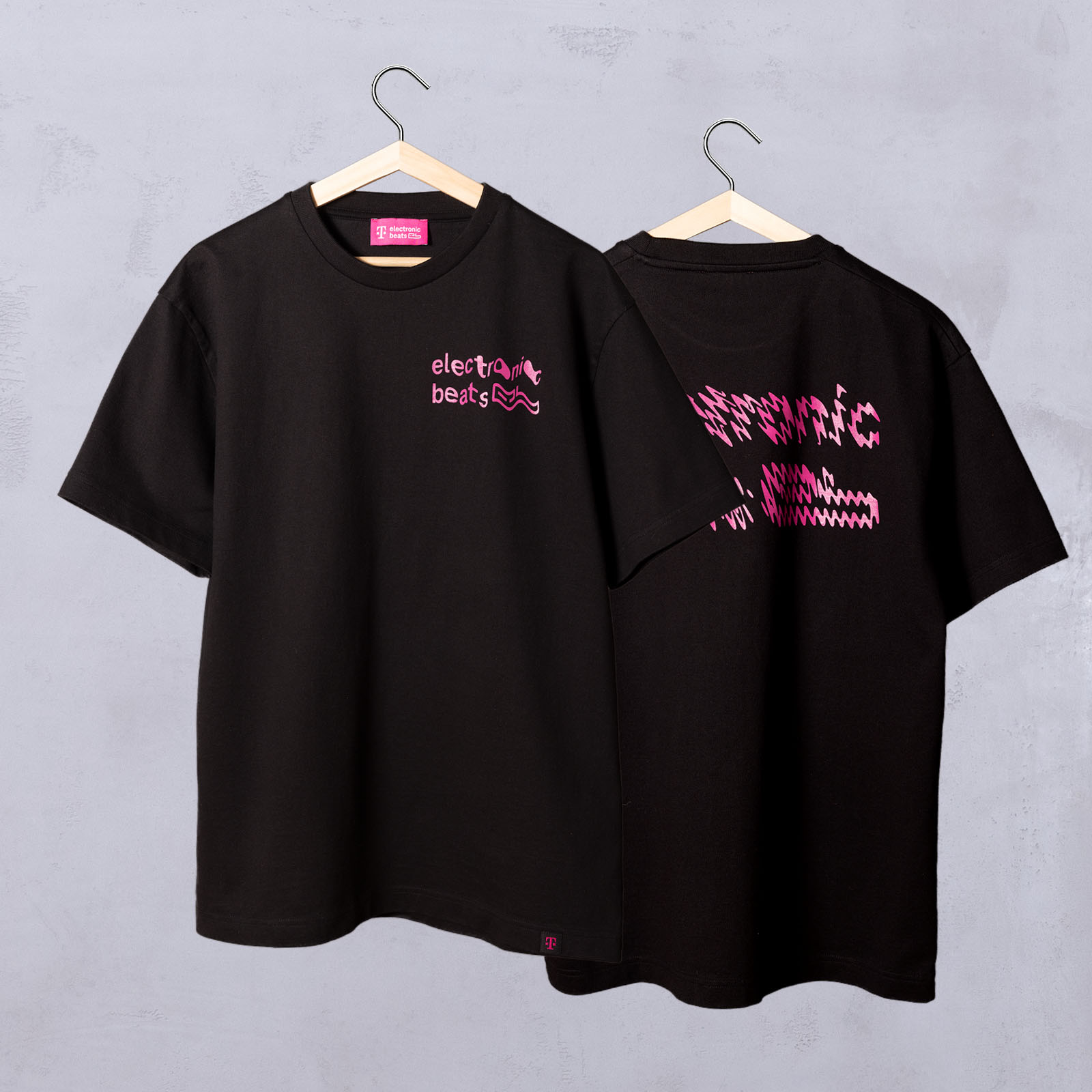 Zwei schwarze T-Shirts an Kleiderbügeln. Auf dem vorderen T-Shirt steht „Electronic Beats“ mit einer rosa Grafik. Das hintere T-Shirt zeigt ein pixeliges rosa-weißes Motiv mit teilweise sichtbarem Text. Beide hängen vor einer schlichten hellgrauen Wand.