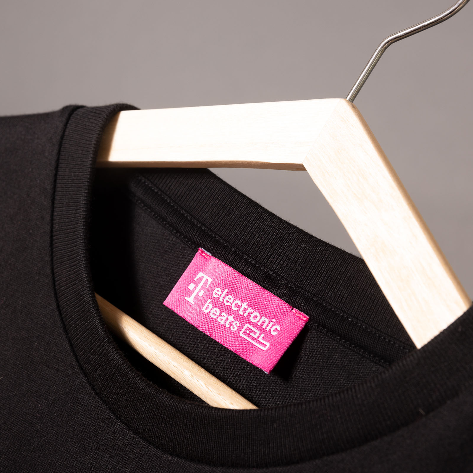 Ein schwarzes T-Shirt auf einem Holzkleiderbügel mit einem rosa Etikett, auf dem in weißer Schrift „electronic beats“ steht. Der Hintergrund ist schlicht und neutral.