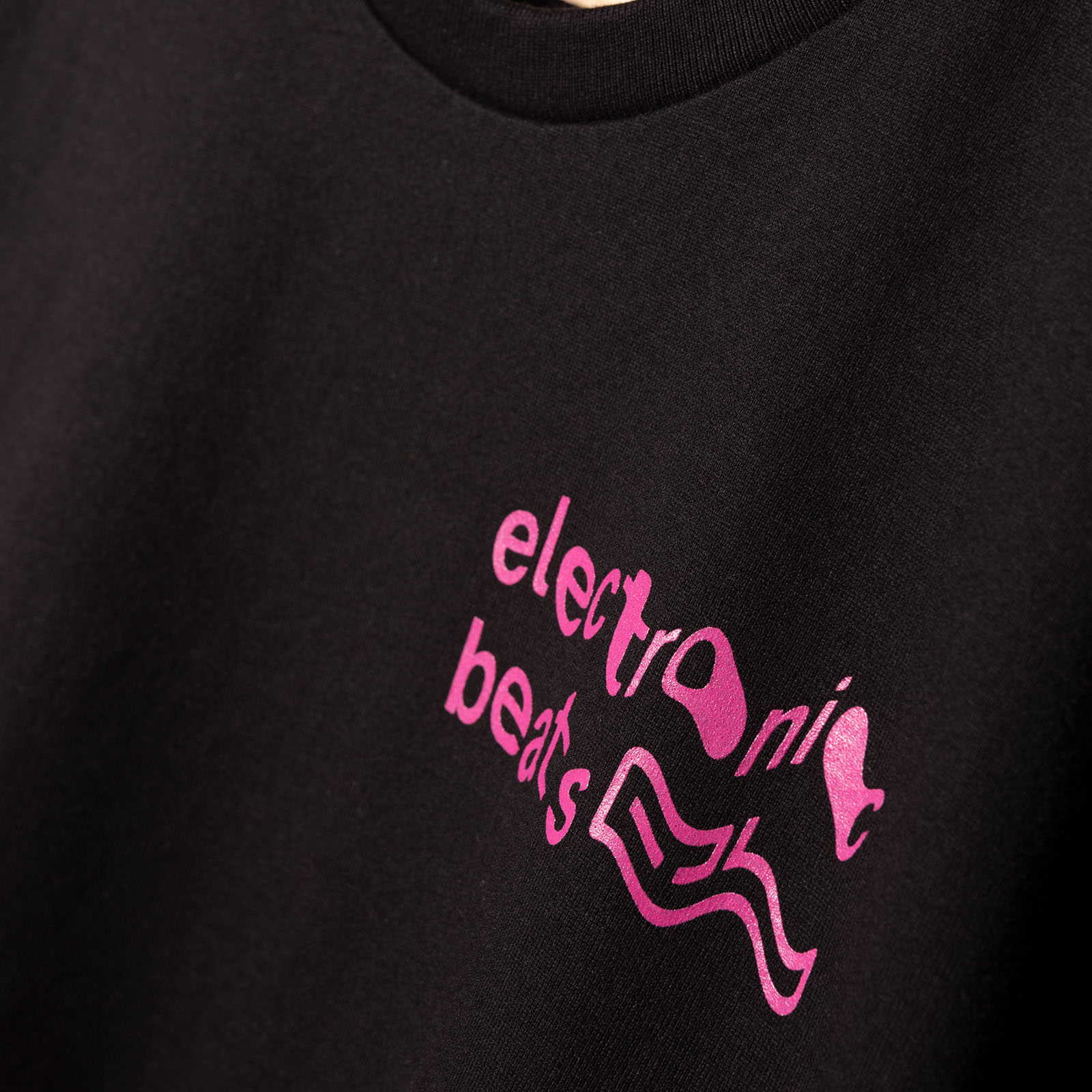 Nahaufnahme eines schwarzen T-Shirts, auf dessen Stoff ein rosa, verzerrter Schriftzug mit der Aufschrift „electronic beats“ sowie abstrakte, wellenförmige Linien aufgedruckt sind.