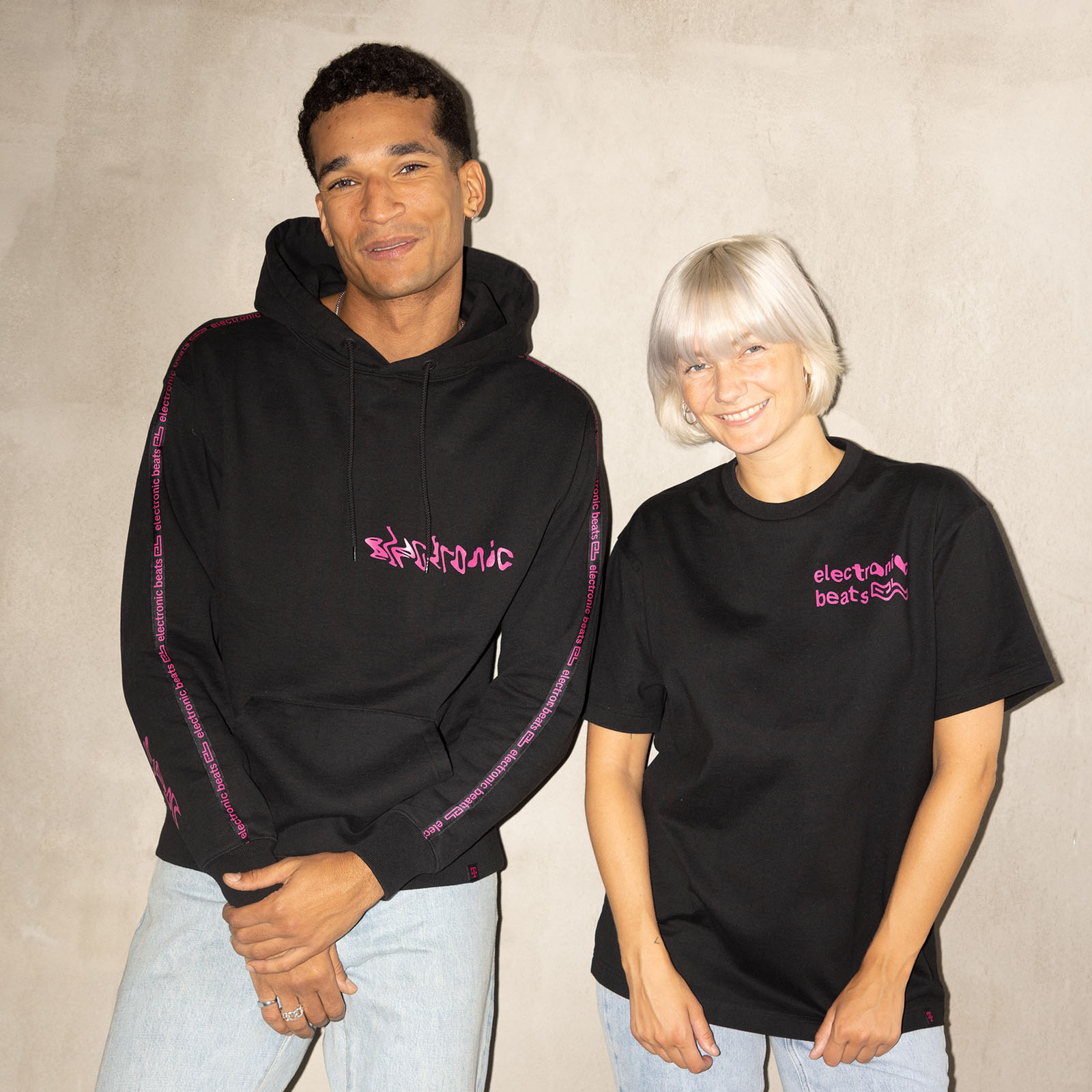 Zwei Personen stehen vor einer beigen Wand. Die eine trägt einen schwarzen Kapuzenpulli mit rosa Aufdruck, die andere ein schwarzes T-Shirt mit rosa Motiven. Beide lächeln und tragen hellblaue Jeans.