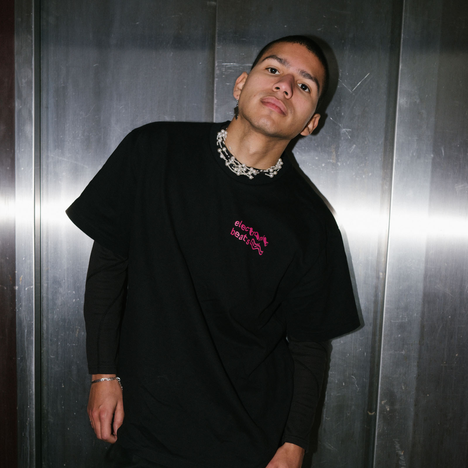 Eine Person, die ein schwarzes T-Shirt und eine gemusterte Halskette trägt, beugt sich leicht zur Seite und posiert mit ausdruckslosem Gesicht vor einem metallischen Hintergrund.