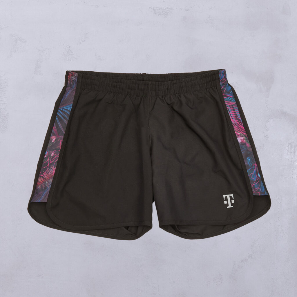 Schwarze Sportshorts mit einem bunten, abstrakten Muster an den Seitenteilen und einem weißen T-Logo am linken Unterschenkel, flach auf einer leicht strukturierten Oberfläche liegend.