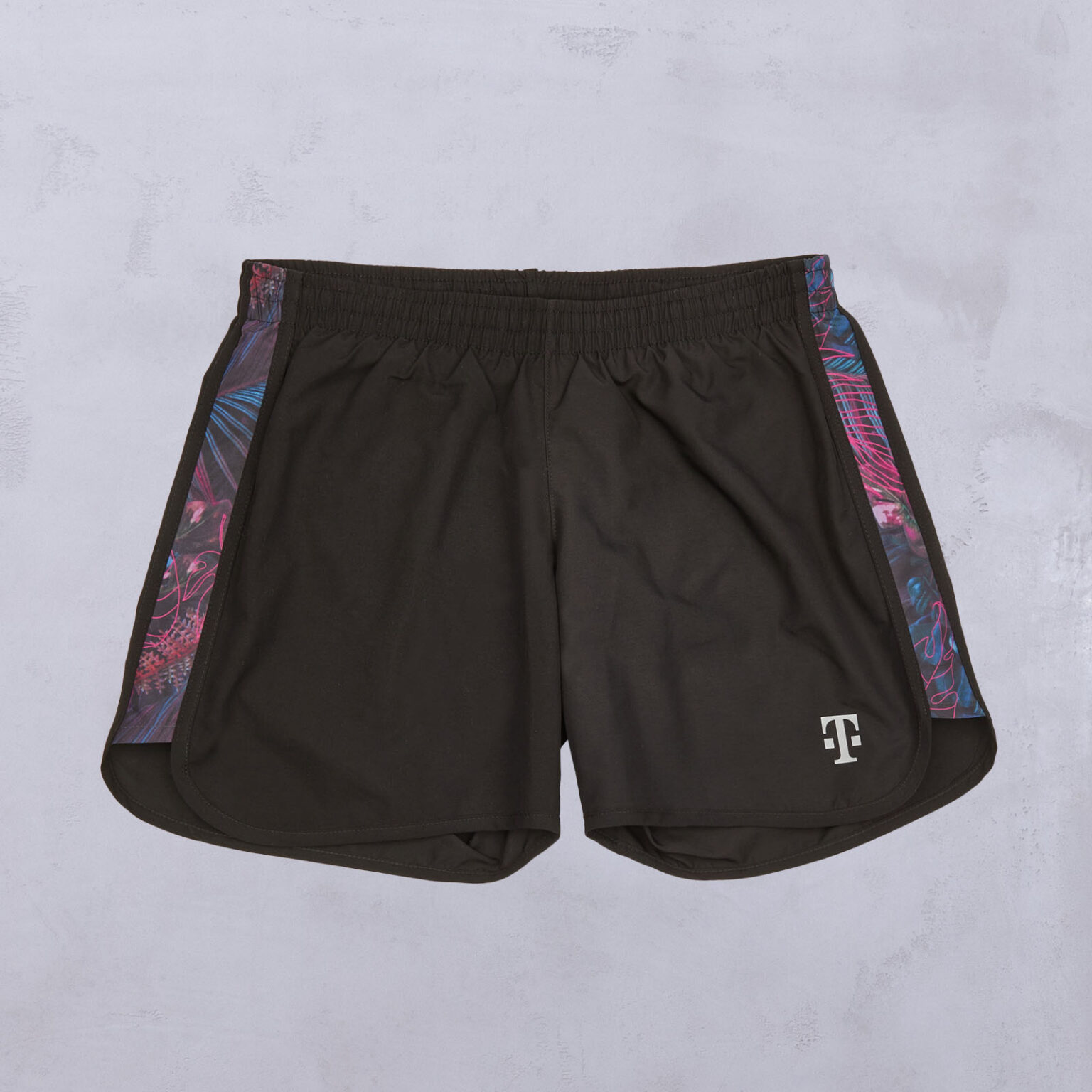 Schwarze Sportshorts mit einem bunten, abstrakten Muster an den Seitenteilen und einem weißen T-Logo am linken Unterschenkel, flach auf einer leicht strukturierten Oberfläche liegend.