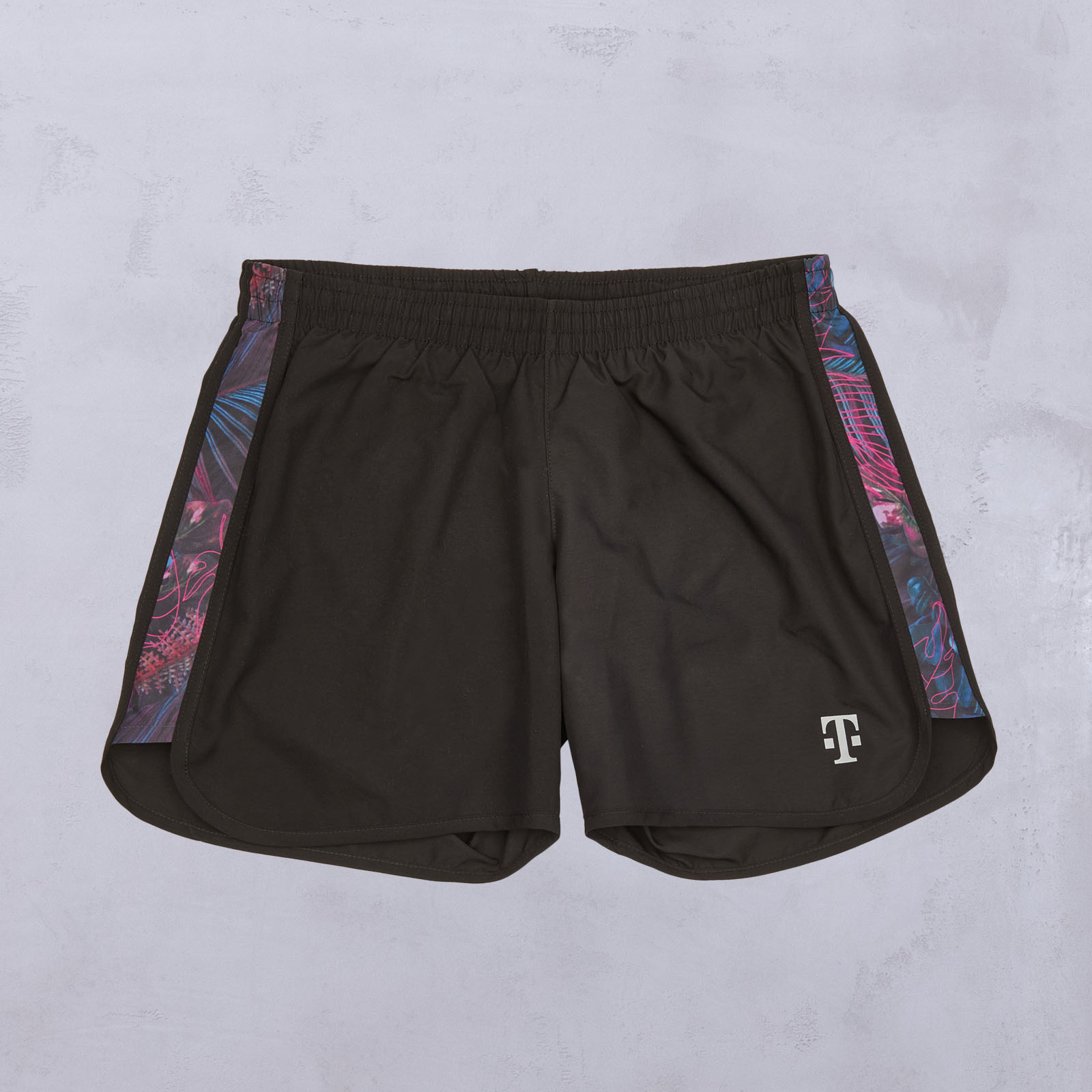 Schwarze Sportshorts mit einem bunten, abstrakten Muster an den Seitenteilen und einem weißen T-Logo am linken Unterschenkel, flach auf einer leicht strukturierten Oberfläche liegend.