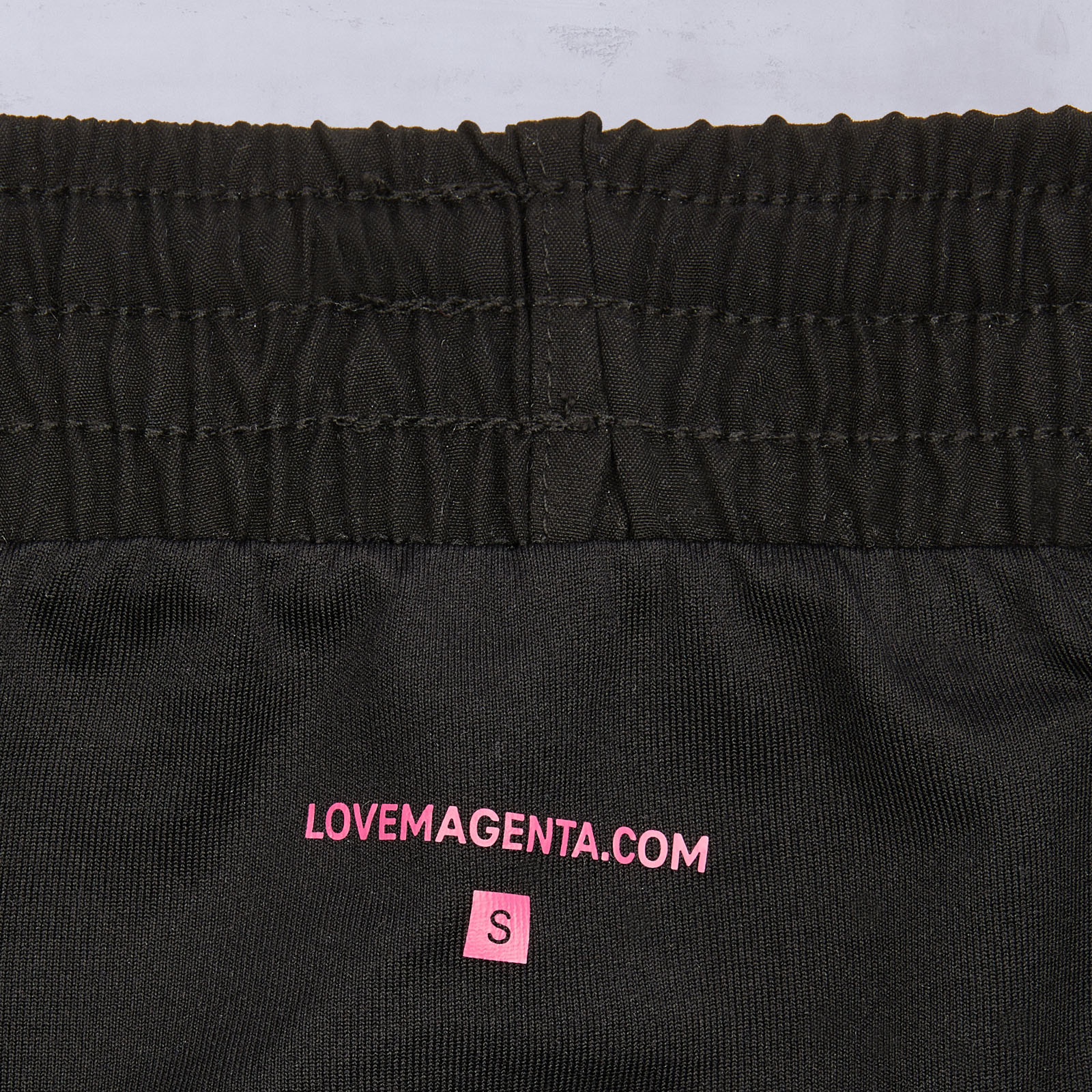 Nahaufnahme eines schwarzen, elastischen Bunds mit dem Aufdruck „LOVEMAGENTA.COM“ in Rosa und einem kleinen rosa Etikett mit dem Buchstaben „S“ darunter, das die Größe „Small“ angibt.