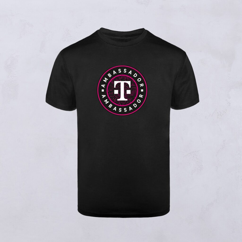 Schwarzes T-Shirt mit einem runden Logo in der Mitte. Das Logo zeigt ein weißes „T“ in einem kastanienbraunen Kreis, umrandet von dem Wort „AMBASSADOR“, das in weißen Buchstaben um den Rand herum wiederholt wird – ein Markenzeichen von Ambassador Designs.
