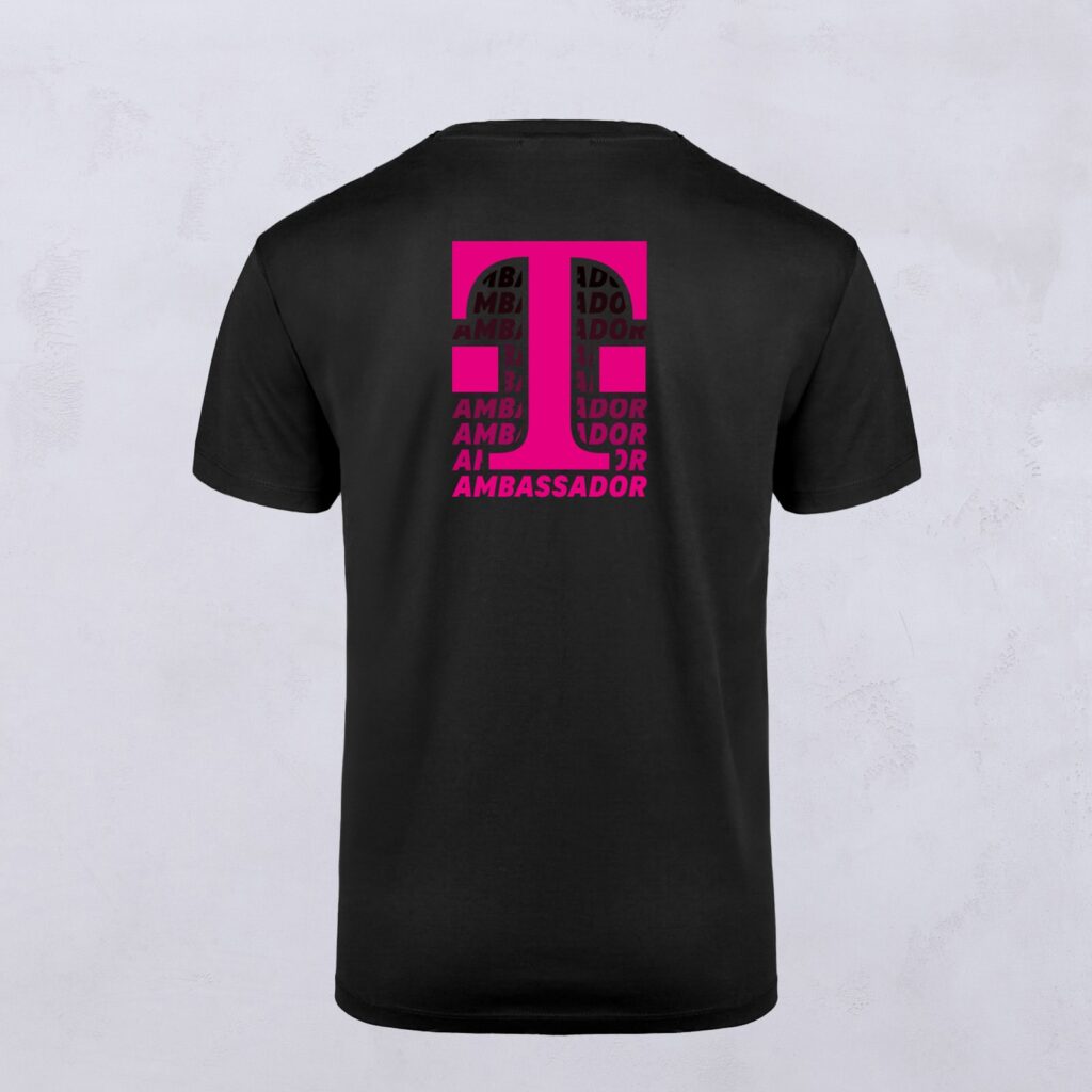 Schwarzes T-Shirt mit einem auffälligen pinkfarbenen KI-Motiv auf der Rückseite, entworfen von LOVE MAGENTA Studio. Das Wort „AMBASSADOR“ wiederholt sich mehrfach innerhalb der Buchstabenformen. Das Shirt wird vor einem einfarbigen hellen Hintergrund präsentiert.