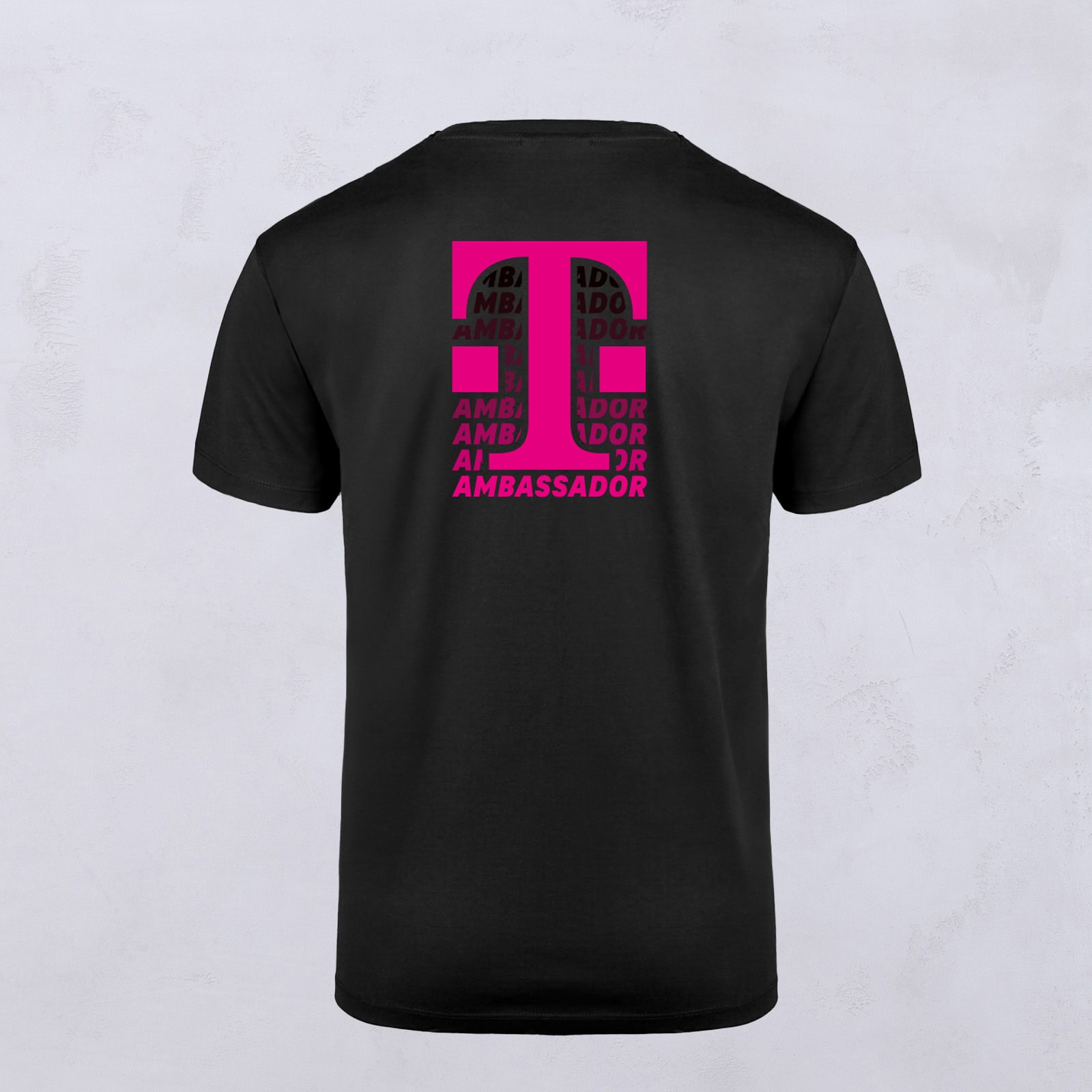 Schwarzes T-Shirt mit einem auffälligen pinkfarbenen KI-Motiv auf der Rückseite, entworfen von LOVE MAGENTA Studio. Das Wort „AMBASSADOR“ wiederholt sich mehrfach innerhalb der Buchstabenformen. Das Shirt wird vor einem einfarbigen hellen Hintergrund präsentiert.