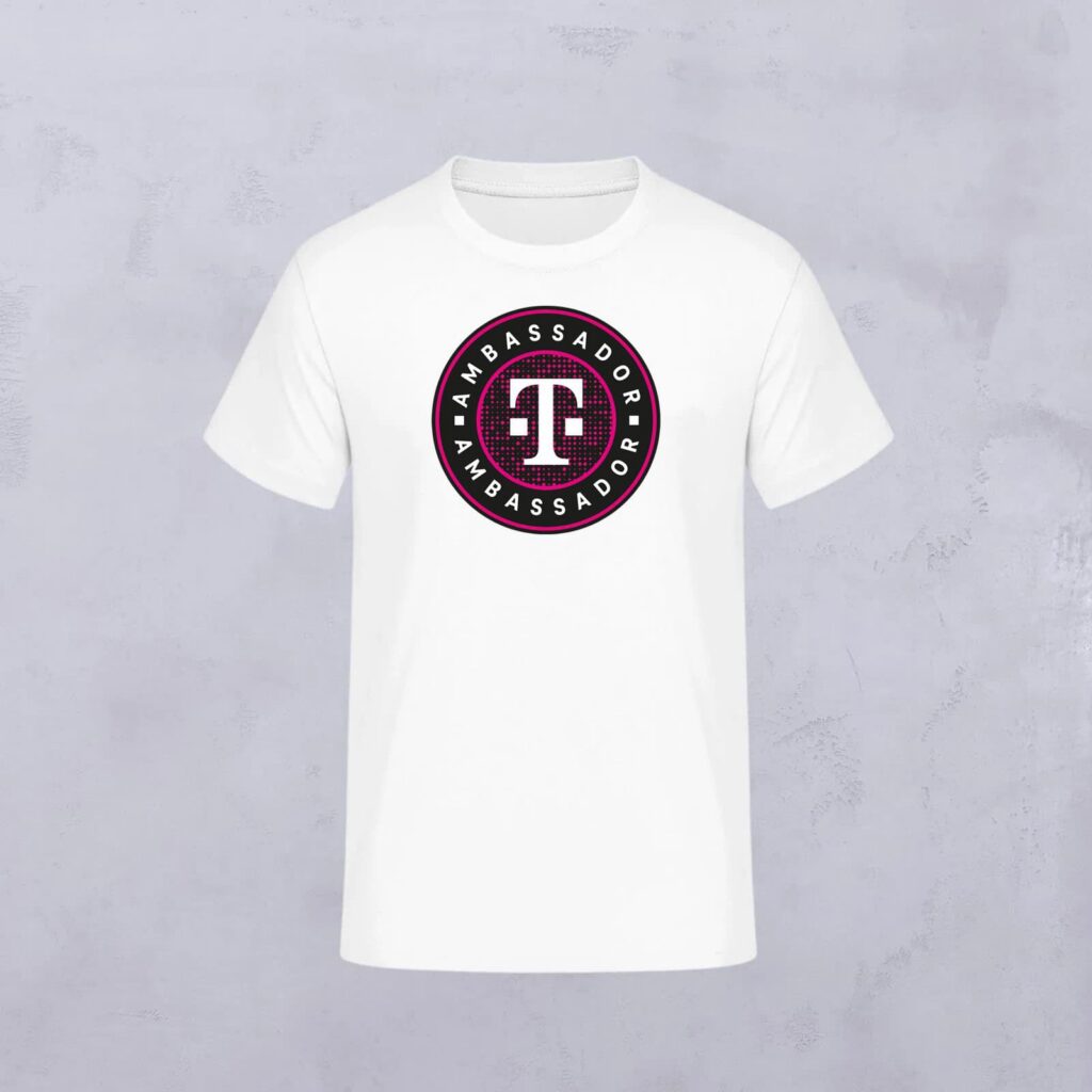 Weißes T-Shirt mit einem runden Logo in der Mitte. Das Logo trägt die Aufschrift „I AM AMBASSADOR“ um ein markantes Symbol herum, das wie eine abstrakte Figur oder ein Buchstabe aussieht, in den Farben Schwarz, Weiß und Rot – ein charakteristisches Detail, das vom LOVE MAGENTA Studio inspiriert ist. Der Hintergrund ist hellgrau.