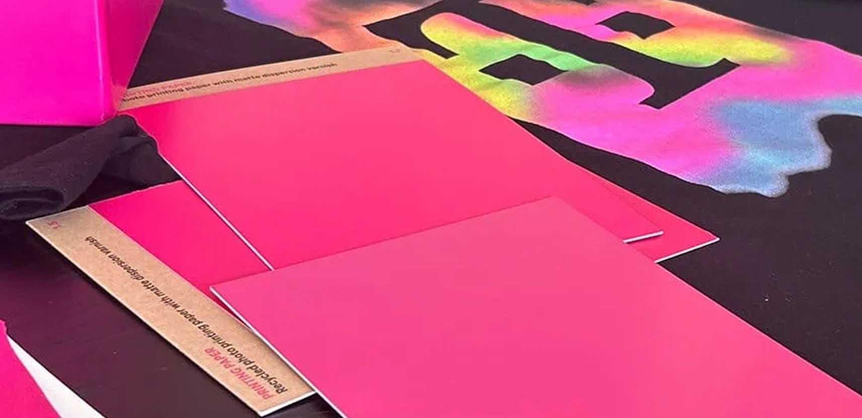 Auf einer Fläche sind mehrere knallrosa Karten und Umschläge angeordnet, im Hintergrund ist ein bunter Batikstoff mit einem großen schwarzen „T“ zu sehen.