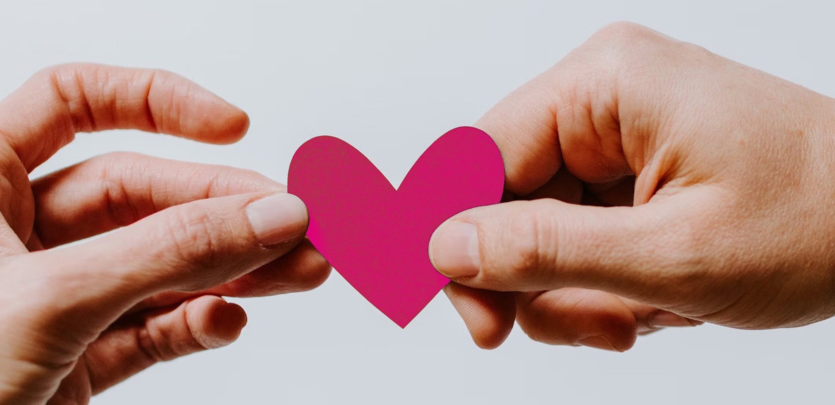 Zwei Hände halten gemeinsam ein rosa Papierherz vor einem hellen, verschwommenen Hintergrund.
