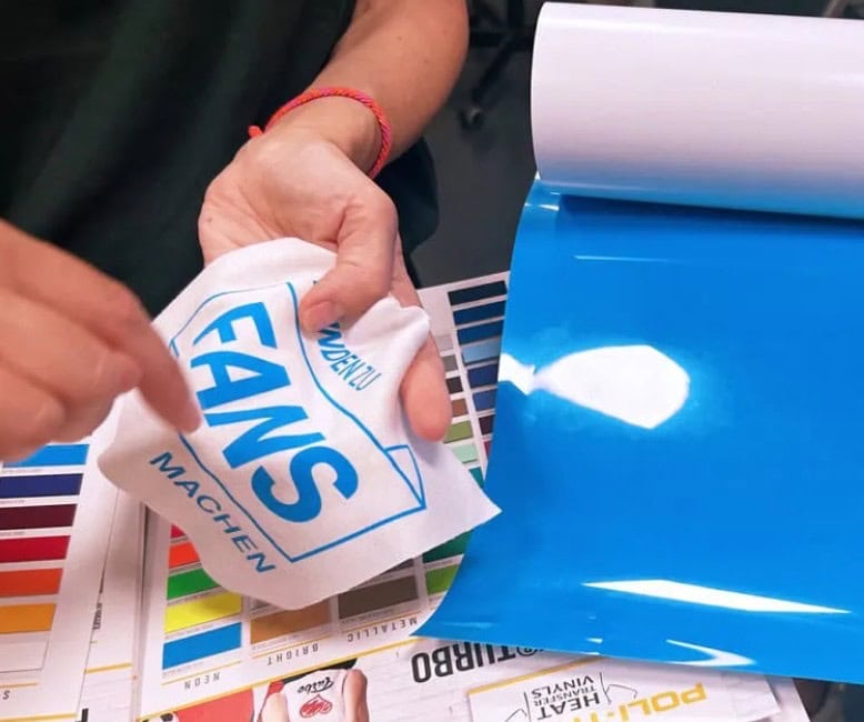 Eine Person zieht ein bedrucktes „FANS MACHEN“-Motiv vom Trägerpapier ab und bereitet sich darauf vor, es per Thermotransfer auf Stoff zu übertragen; daneben liegen ein blaues Vinylblatt und Farbmuster.