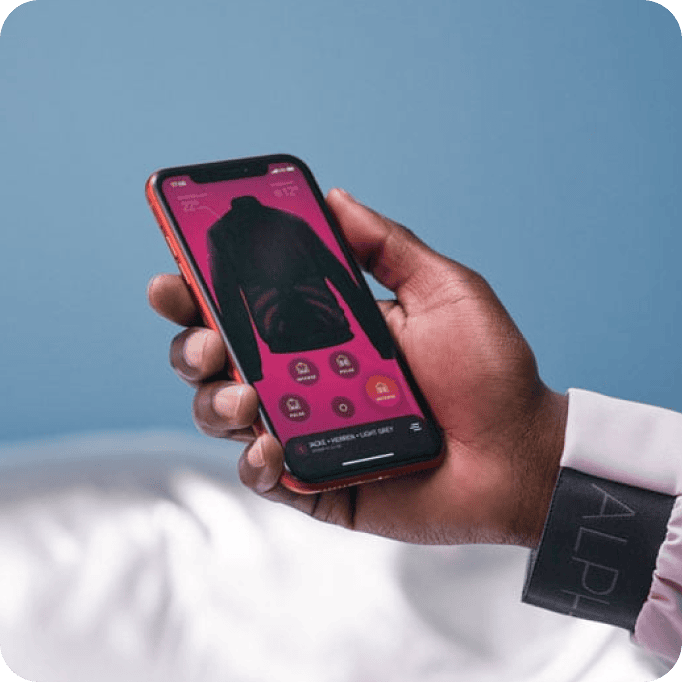 Eine Person hält ein Smartphone in der Hand, auf dem eine Shopping-App angezeigt wird. Auf dem Bildschirm ist ein schwarzer Kapuzenpulli vor einem rosa Hintergrund zu sehen, daneben befinden sich verschiedene Optionsschaltflächen. Auf dem Ärmel der Person steht „ALPHA“.