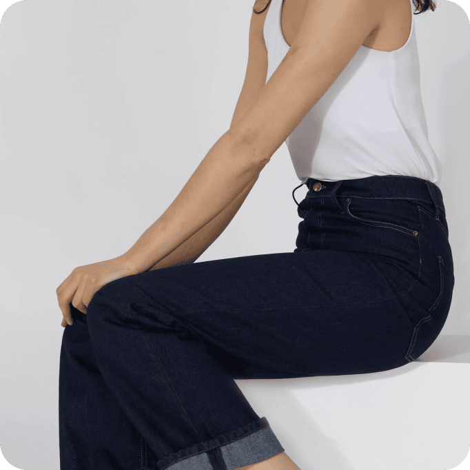 Eine Person, die ein weißes, ärmelloses Oberteil und dunkelblaue Jeans trägt, sitzt auf einer weißen Fläche vor einem hellen Hintergrund. Zu sehen sind nur der Oberkörper und die Beine; das Gesicht ist nicht zu erkennen.
